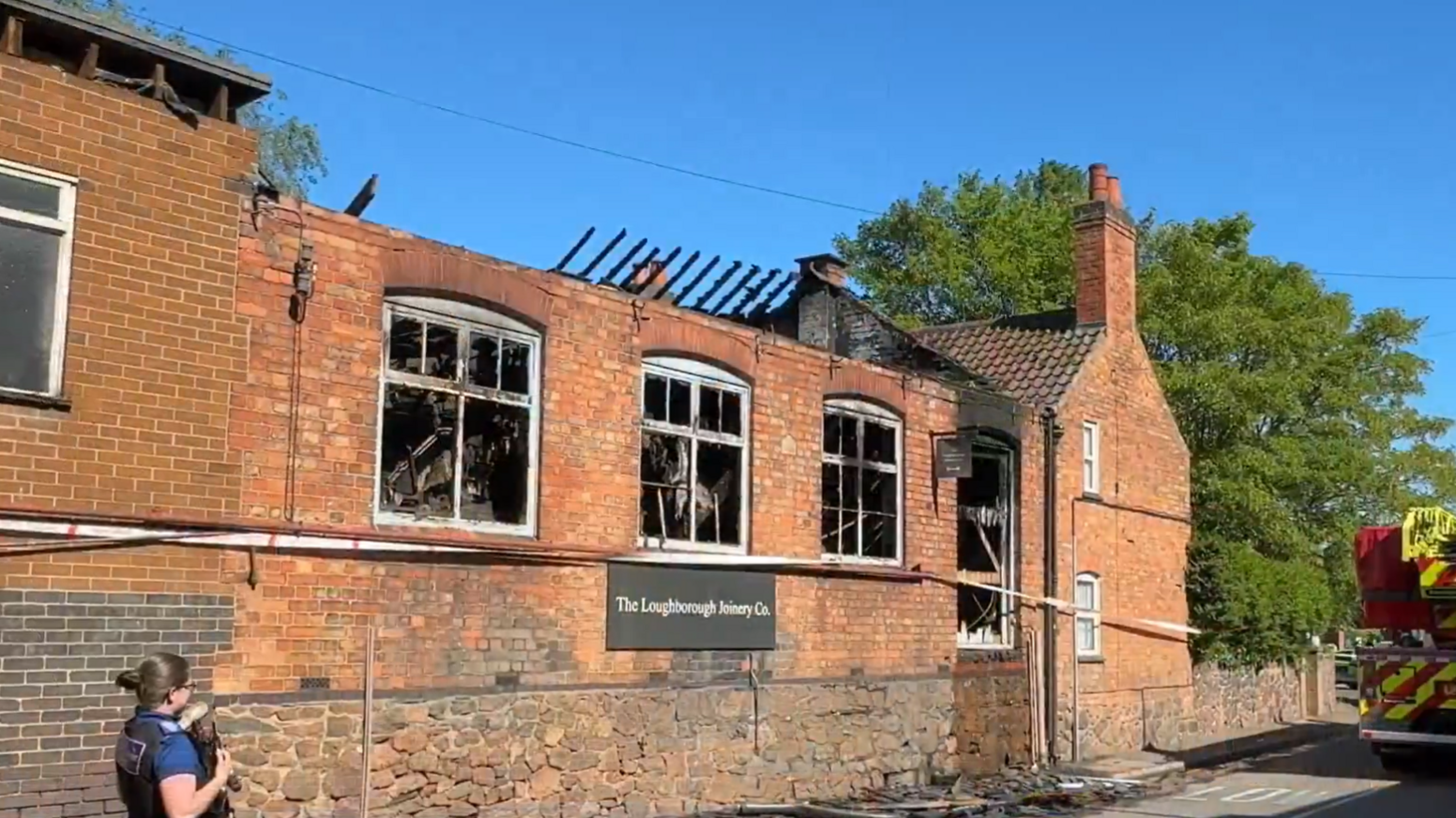 A brick building with extensive damage to its roof and windows. A police officer stands outside and a fire engine can be seen