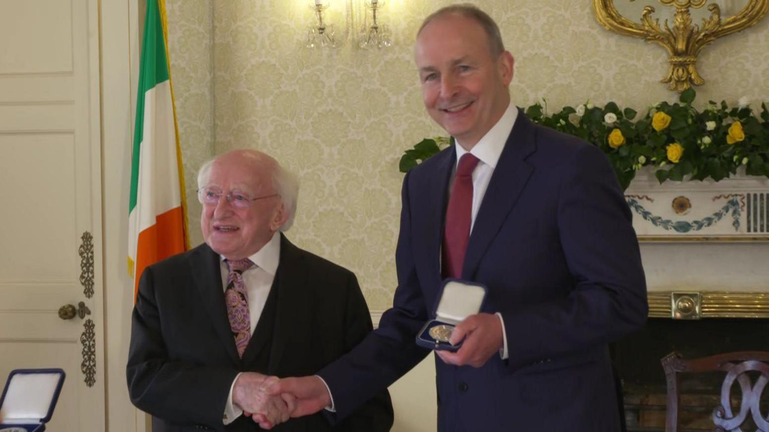 Michael D Higgins - a shorter man with white hair and glasses who is wearing a dark suit jacket, a patterned tie and a white collared shirt shakes the hand of Micheál Martin, who is wearing a navy suit jacket, a white collared shirt and a red tie. They are standing in a well decorated state room with a Irish tricolour in the background.