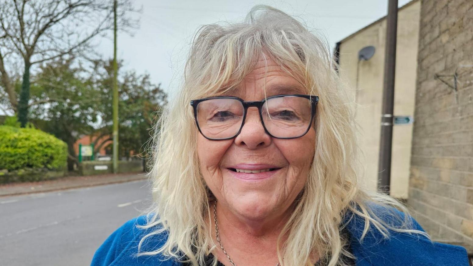 Woman with blonde hair and a fringe looks directly into the camera and smiles. she is wearing black square glasses and a blue coat.