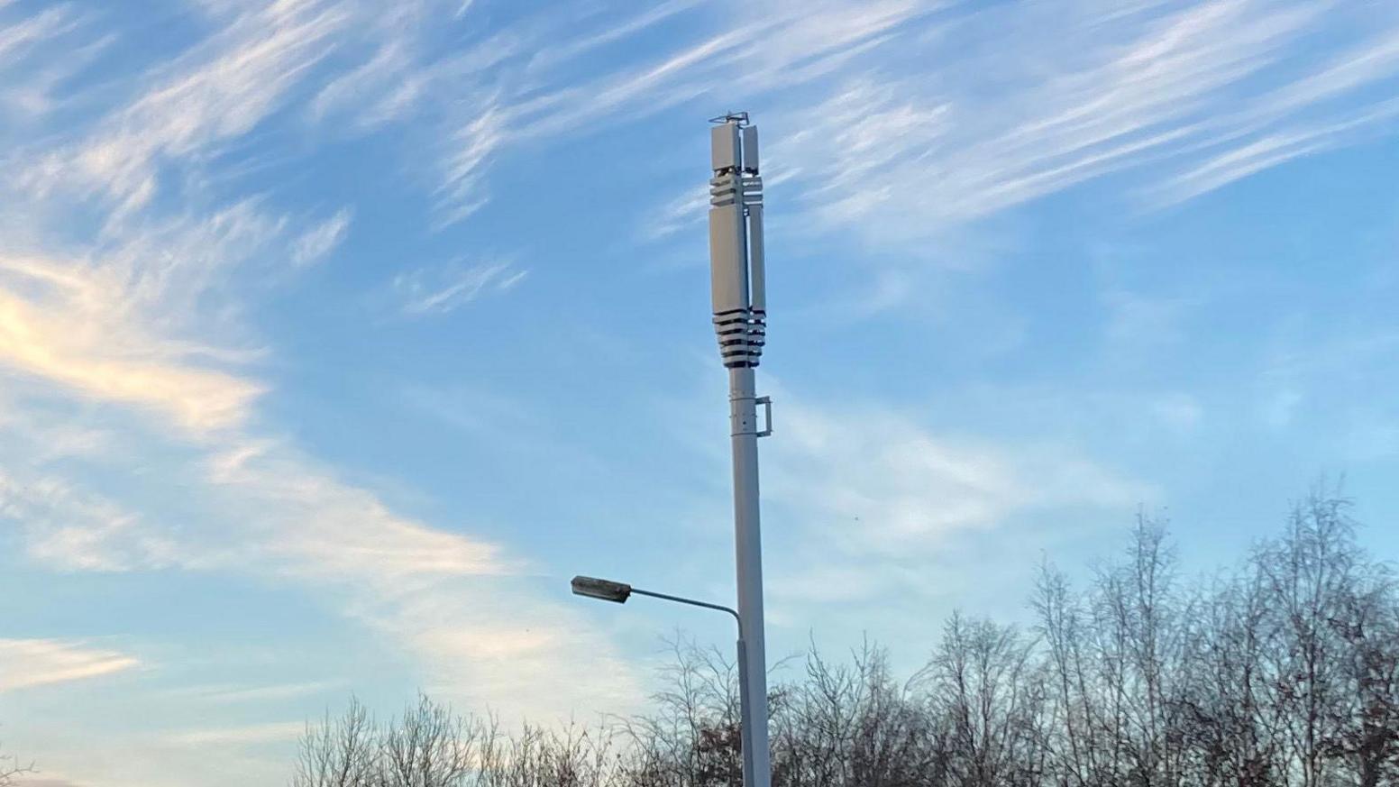 A 5G mast pole with blue skies, clouds and trees behind it.
