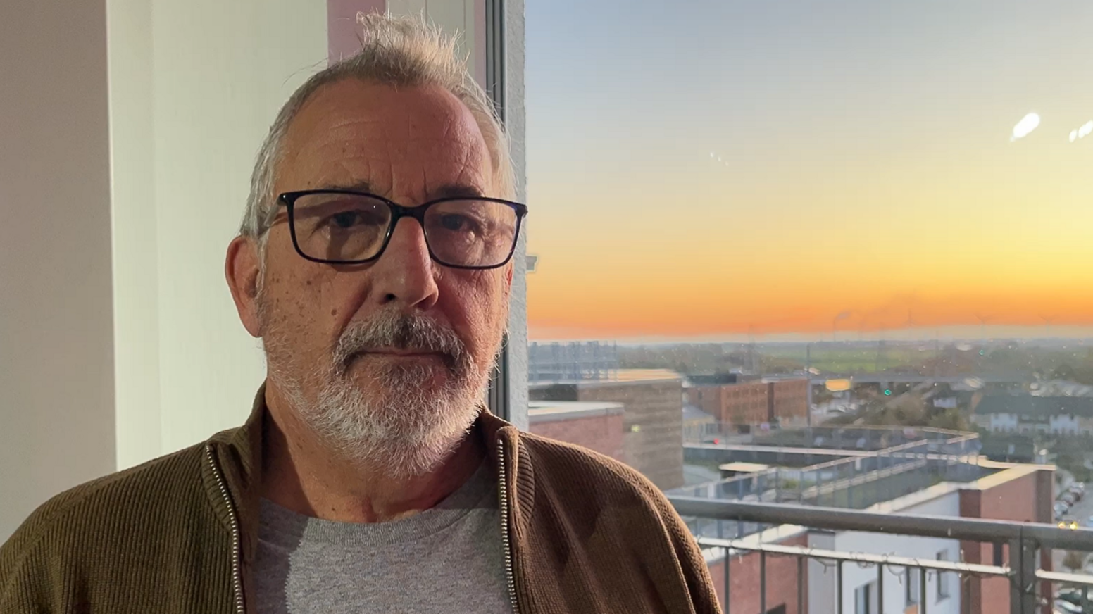 Michael Ferguson wears square framed glasses and has grey hair and a beard. He is inside his flat with sunset behind him outside the window.