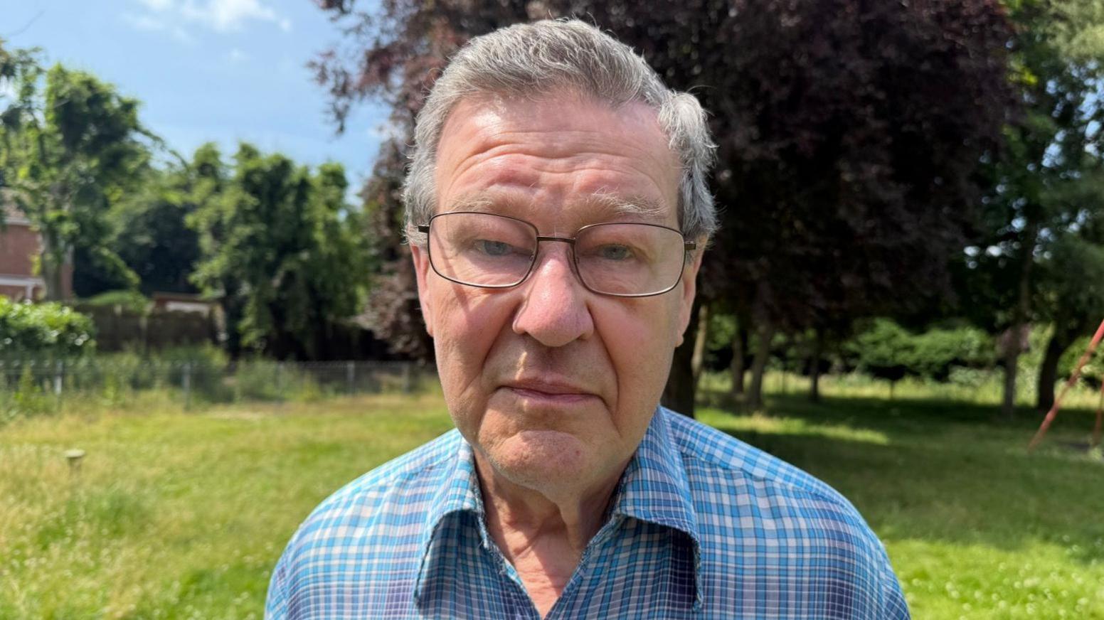 A man is looking at the camera with a neutral expression. He has grey hair, and is wearing glasses. He's also wearing a blue and white checked shirt. In the background there are lots of trees and long grass.