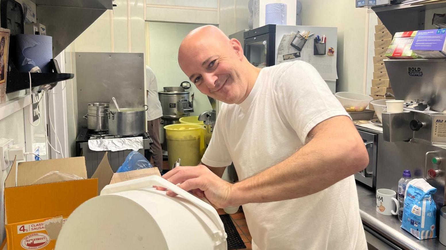 Ronnie Christodoulou working at the back of his chip shop. He is wearing a white t-shirt and is bald. 