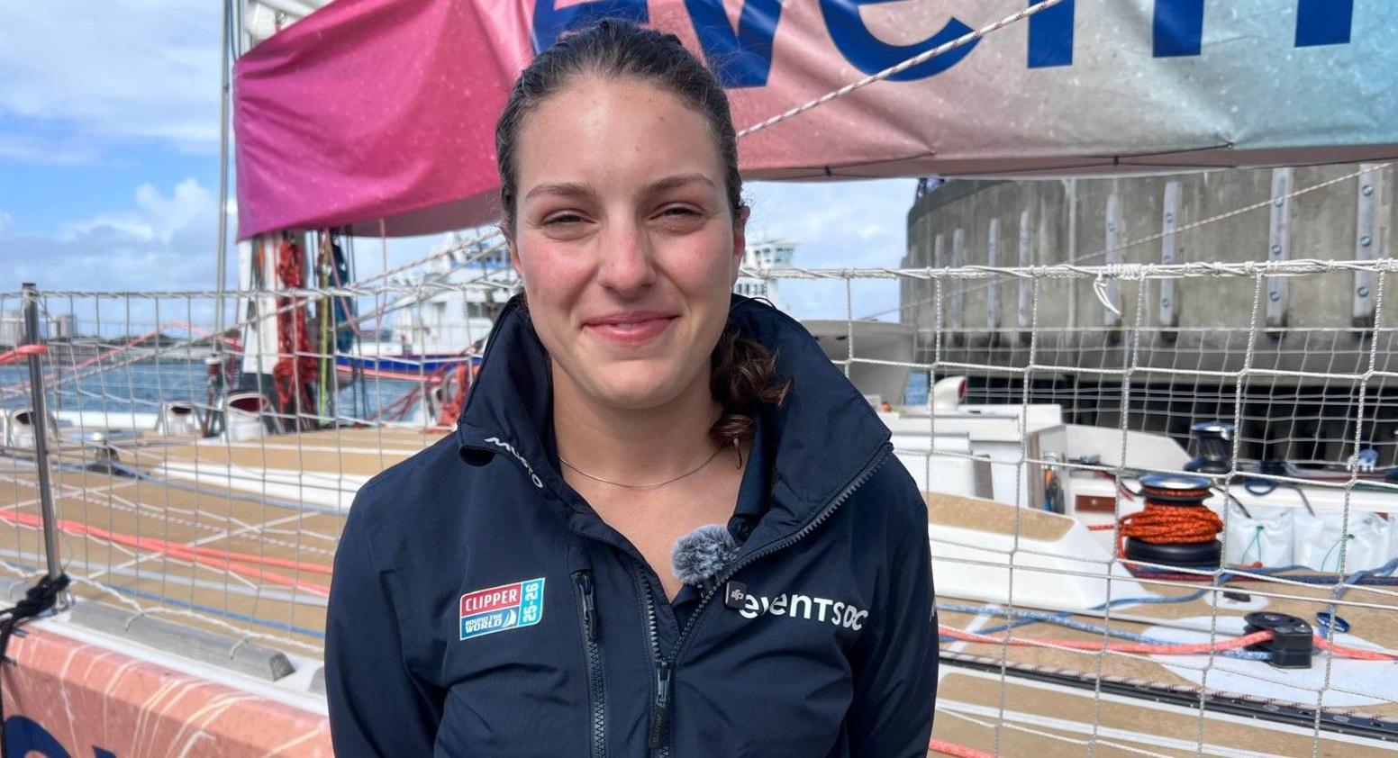 Ella Hebron smiling for the camera. She is standing on the quayside with one of the yachts behind her. She has tied back brown hair and is wearing a navy blue sailing jacket.