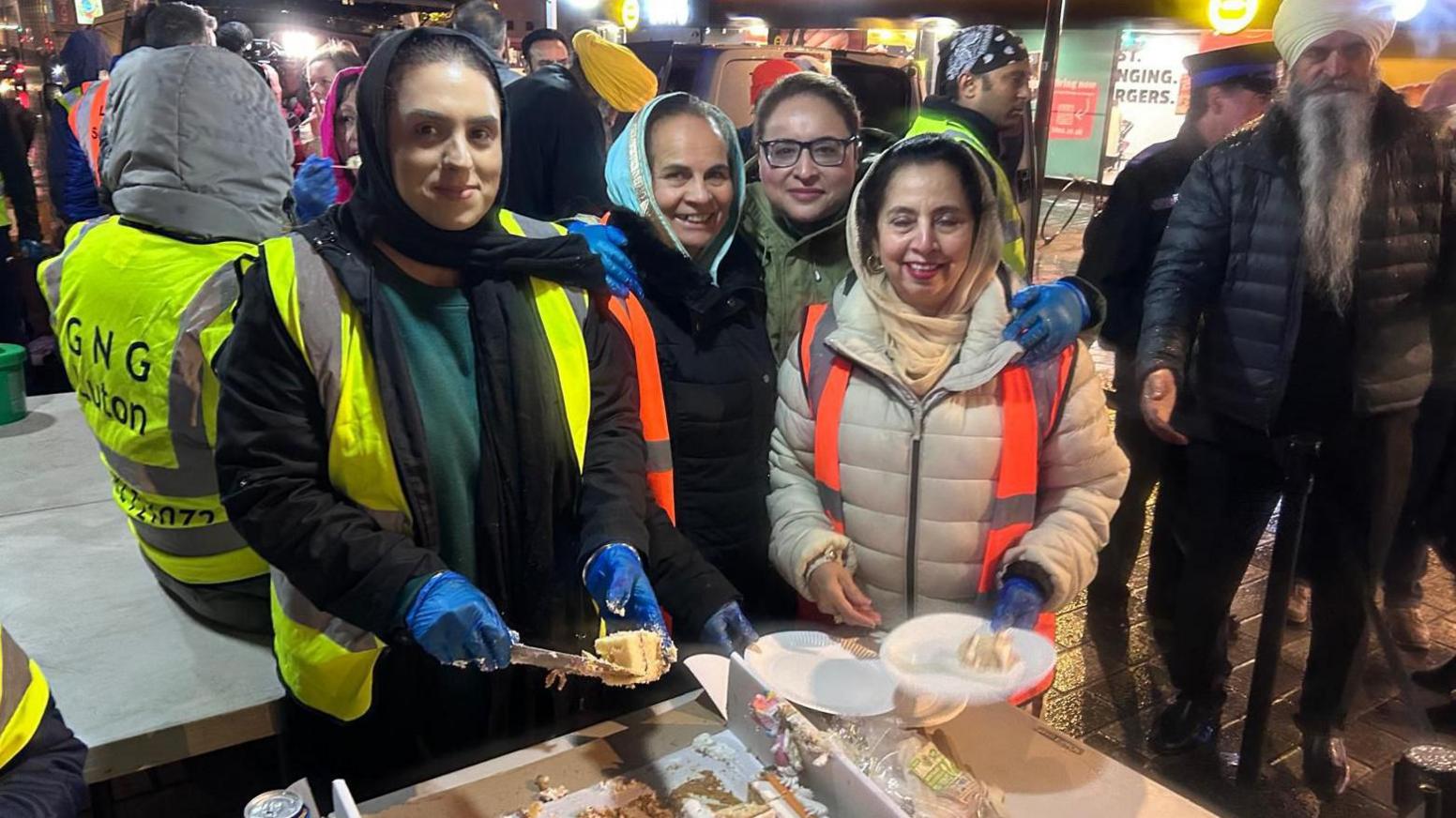 A group of women smile at the camera as they stand outside at the Luton Sikh Soup Kitchen. They wear coats, blue plastic gloves and high vis jackets, and two of them wear headscarves. They are serving up cake onto white paper plates. In the background a Sikh man with a long grey beard and a white turban also looks at the camera. In the background are other volunteers and a van with its back door open.