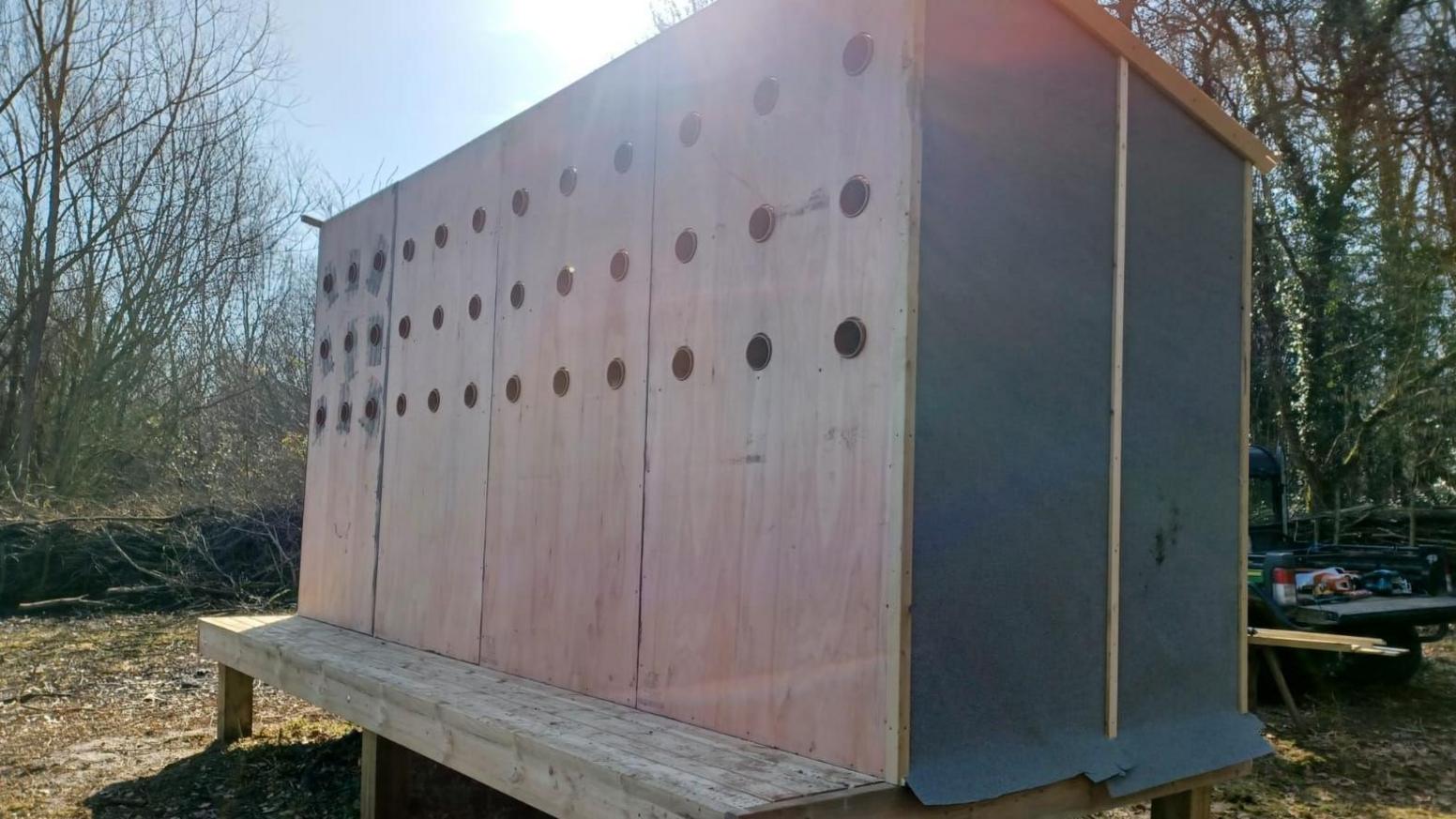 A tall wooden box with three rows of 12 circular holes in the top half of the structure.
It is standing on a wooden platform on a dirt surface with trees and scrub in the background.