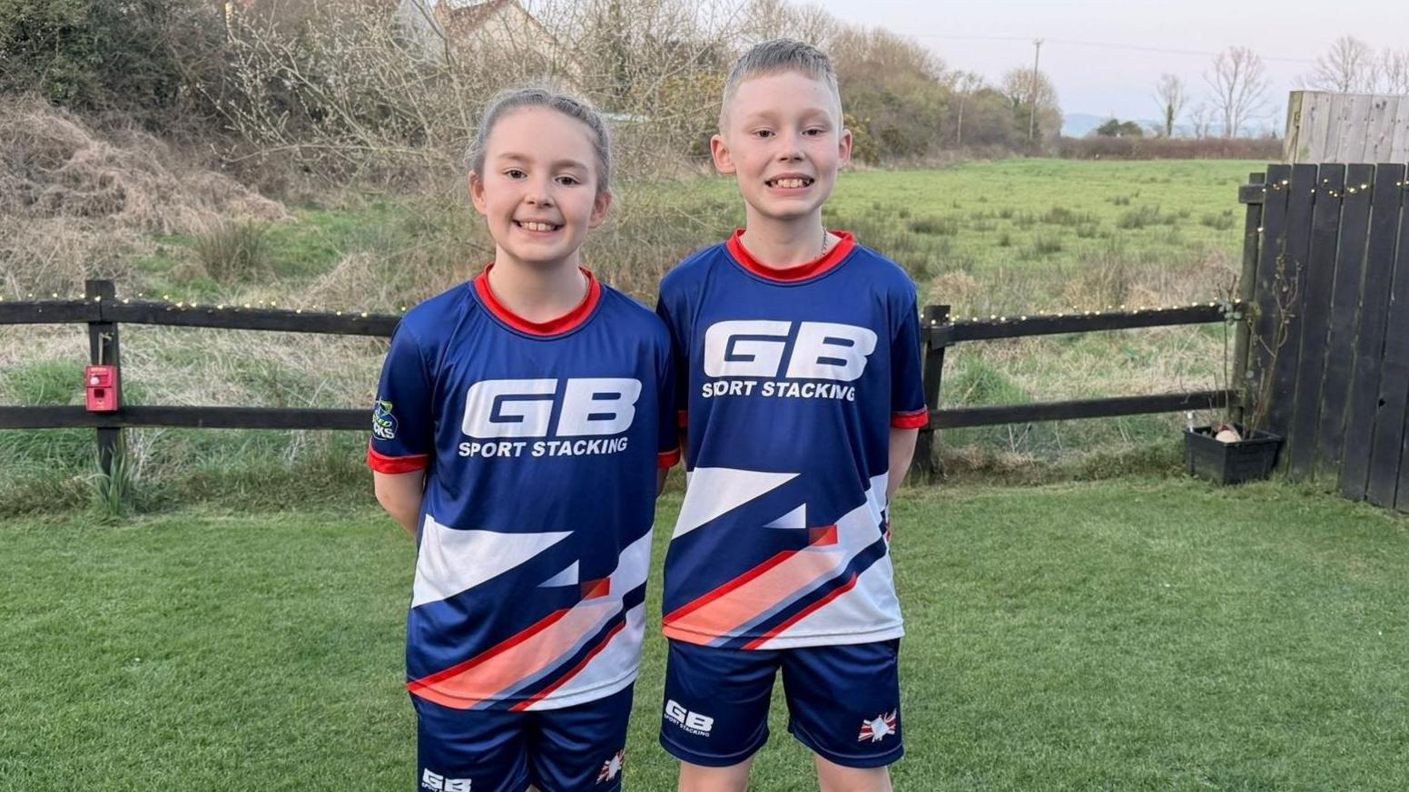 Logan and Ivy are children and are smiling in blue sports uniforms that read "GB sport stacking" on the front in white font. They are standing with their hands behind their backs in a wooden fenced garden with a field in the background. They both have blonde hair, Ivy's is pulled back behind her head, Logan's is short.