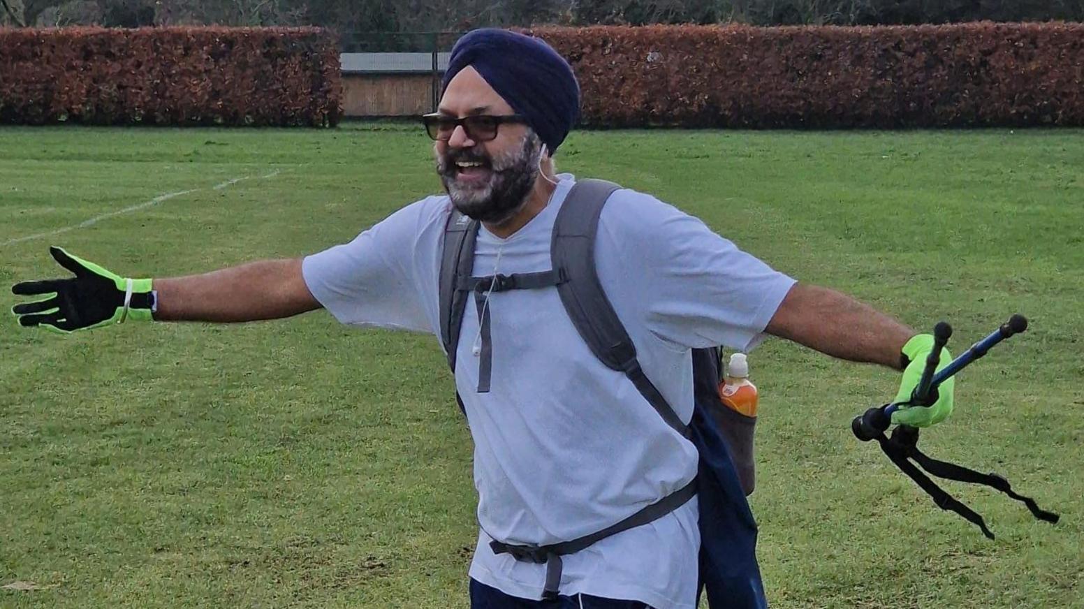 A man with a dark and grey beard wearing a dark turban and a white shirt with a grey backpack. He has his arms stretched out and his is smiling.