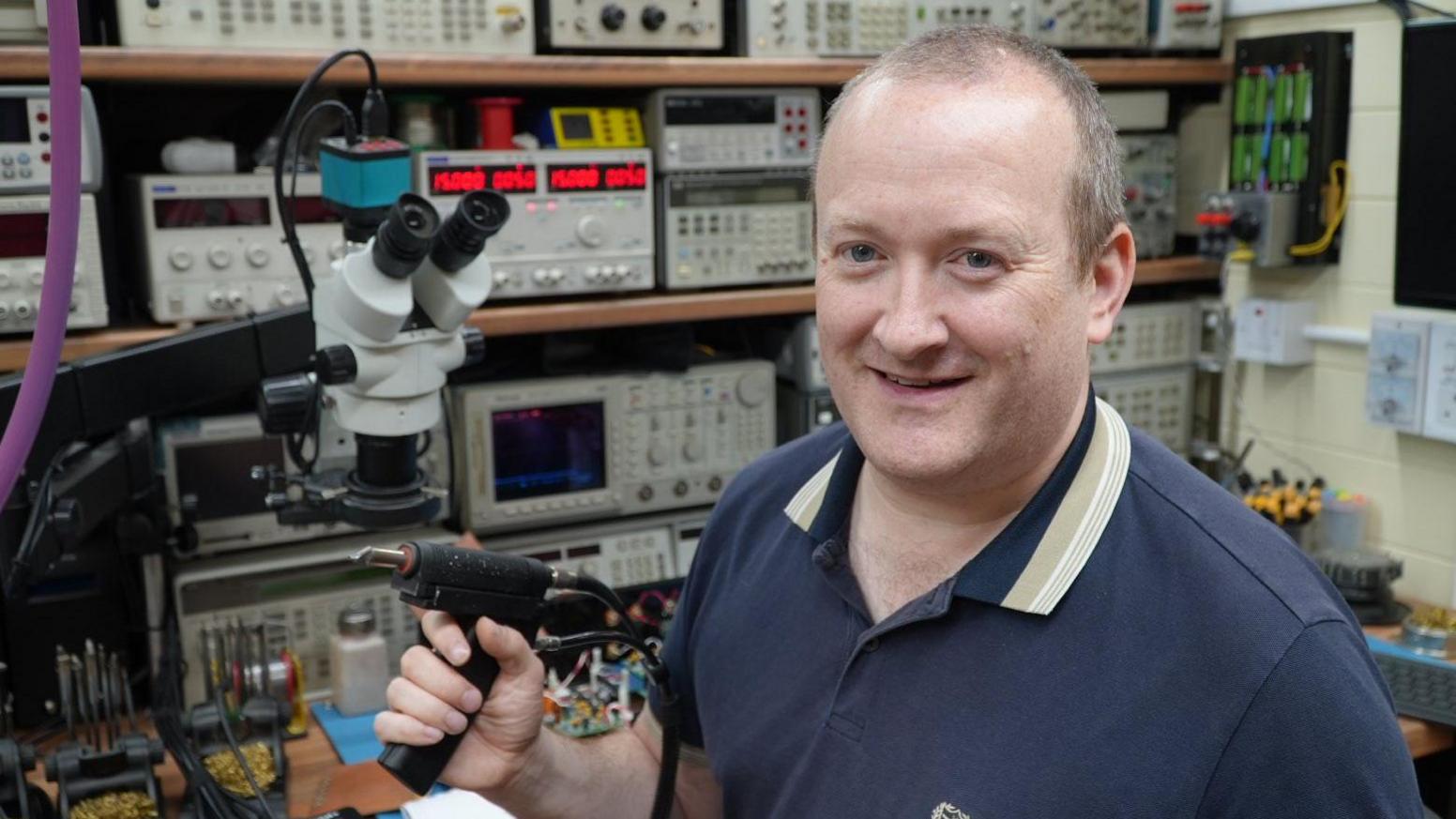 Mark Maher holding a soldering device, behind him are shelves full of electrical equipment