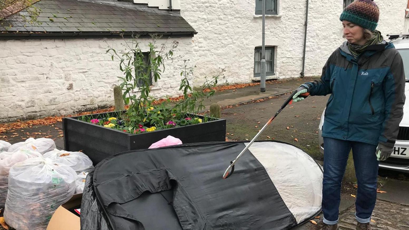 A spray tan booth and lots of full white bin bags in a pile beside a flower bed with a woman holding a litter picker to the right. She is wearing a bobble hat and waterproof jacket and has a look of disgust on her face as she looks at the fly-tipped item