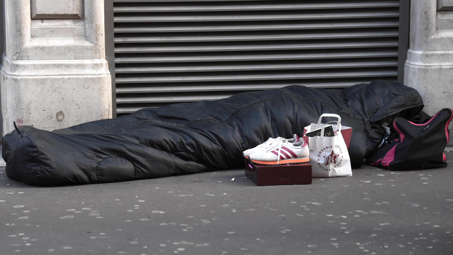 Image of a person sleeping stretched out on a pavement, hidden within a dark sleeping bag with a hood over their head which is resting on a black bag with pink trimming. In front of them is a small Pret paper bag and a pair of white and pink trainers resting on a box