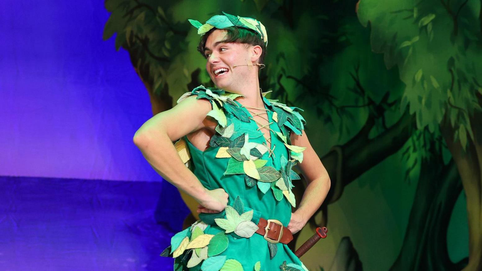 Bradley Riches is dressed in a green tunic and cropped trousers with leaves attached. He is opened mouthed as he is acting in a scene.