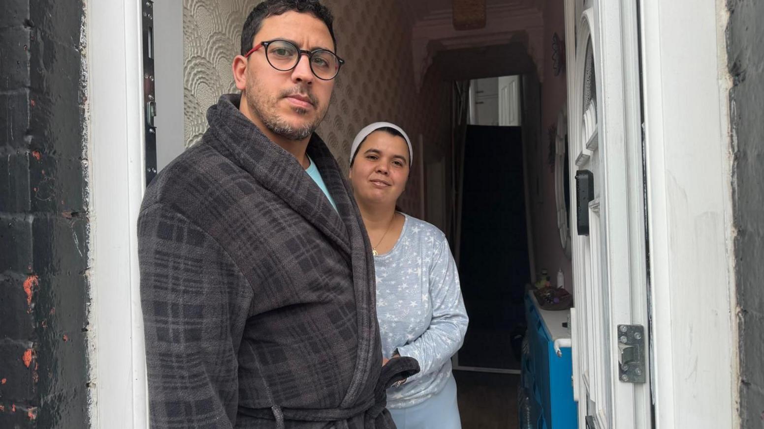 Mohammed Lamaoufi, 42, who has black hair, light stubble, and is wearing a grey cheque patterned dressing gown and glasses, stands in the doorway of a terraced house next to Khadeja Daoud, 34, who wears a light blue top and leggings