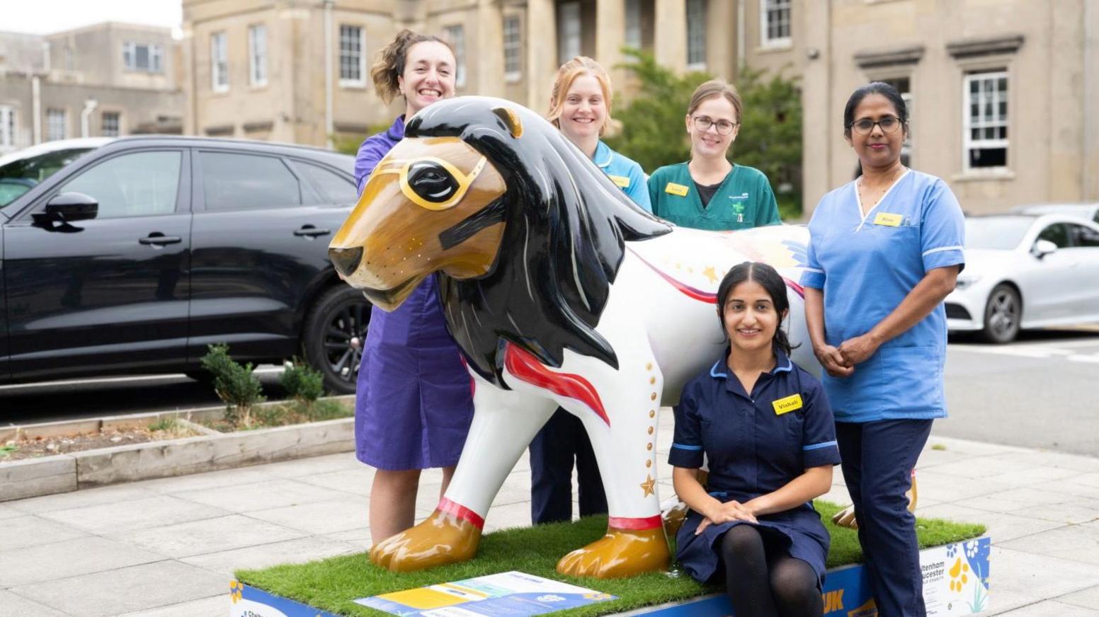 Five oncology staff wearing their uniform and yellow name badges surrounding a lion sculpture. The lion sculpture mimics Elvis Presley with painted sunglasses and white outfit with gold buttons and red stripes down the side.