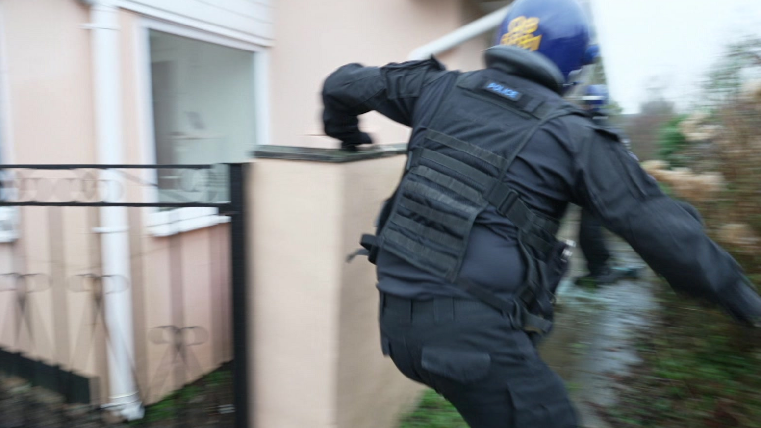 The rear view of a police officer in a blue helmet running around a pale-coloured house.