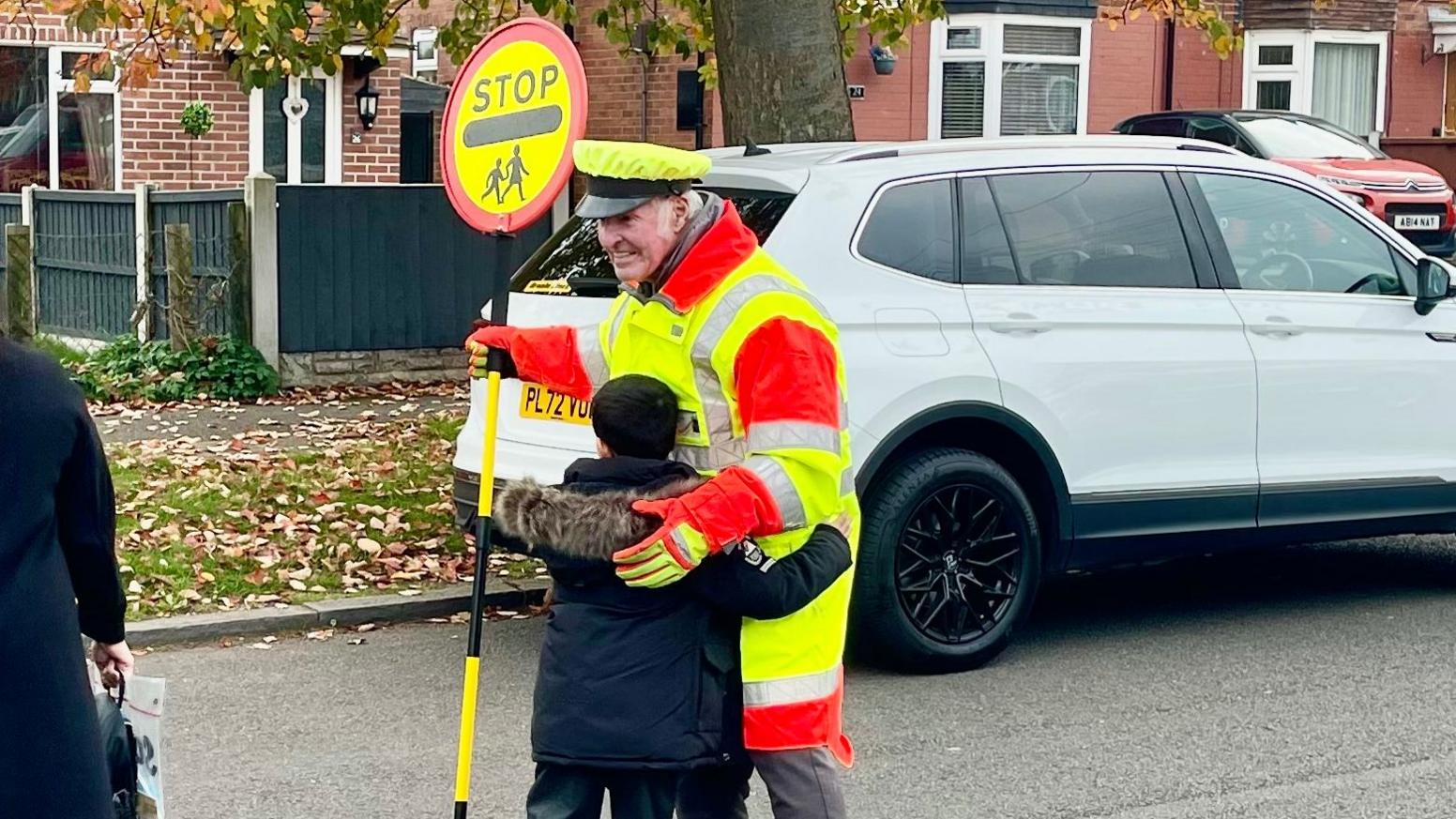 A man stands in the road dressed in a high vis peaked hat, coat and gloves holding a STOP sign. He has white hair and is wearing a grey shirt beneath his uniform. A boy with black hair and a navy parka is giving him a hug. On the other side of the road is a large tree, pavement, grassy areas, semi-detached houses, a white car parked on the pavement and a red car on a driveway.