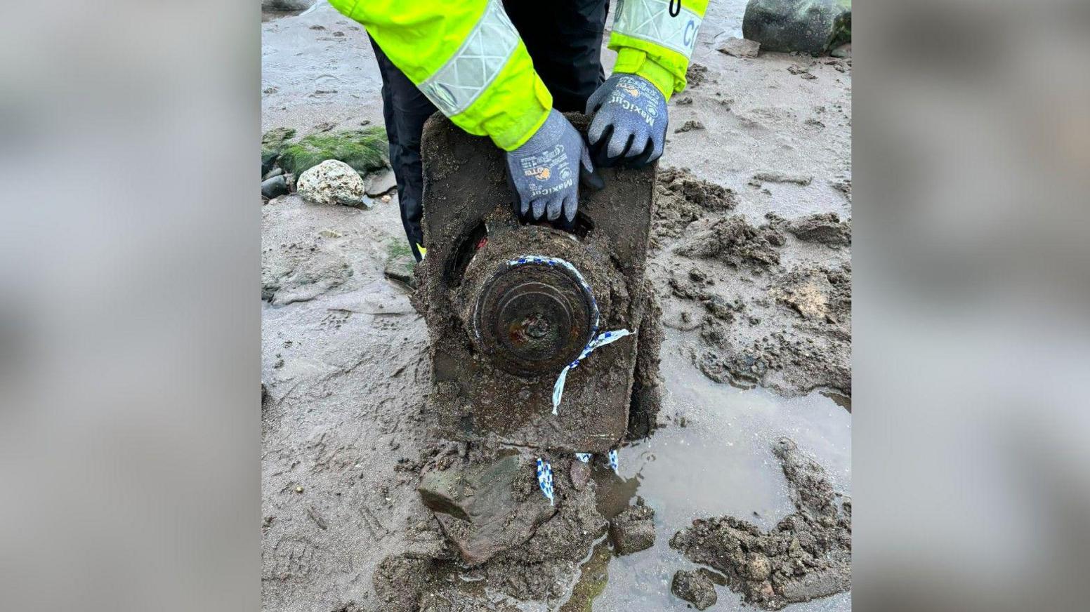 A person on the beach wearing a high-vis jacket and gloves holds the speaker up