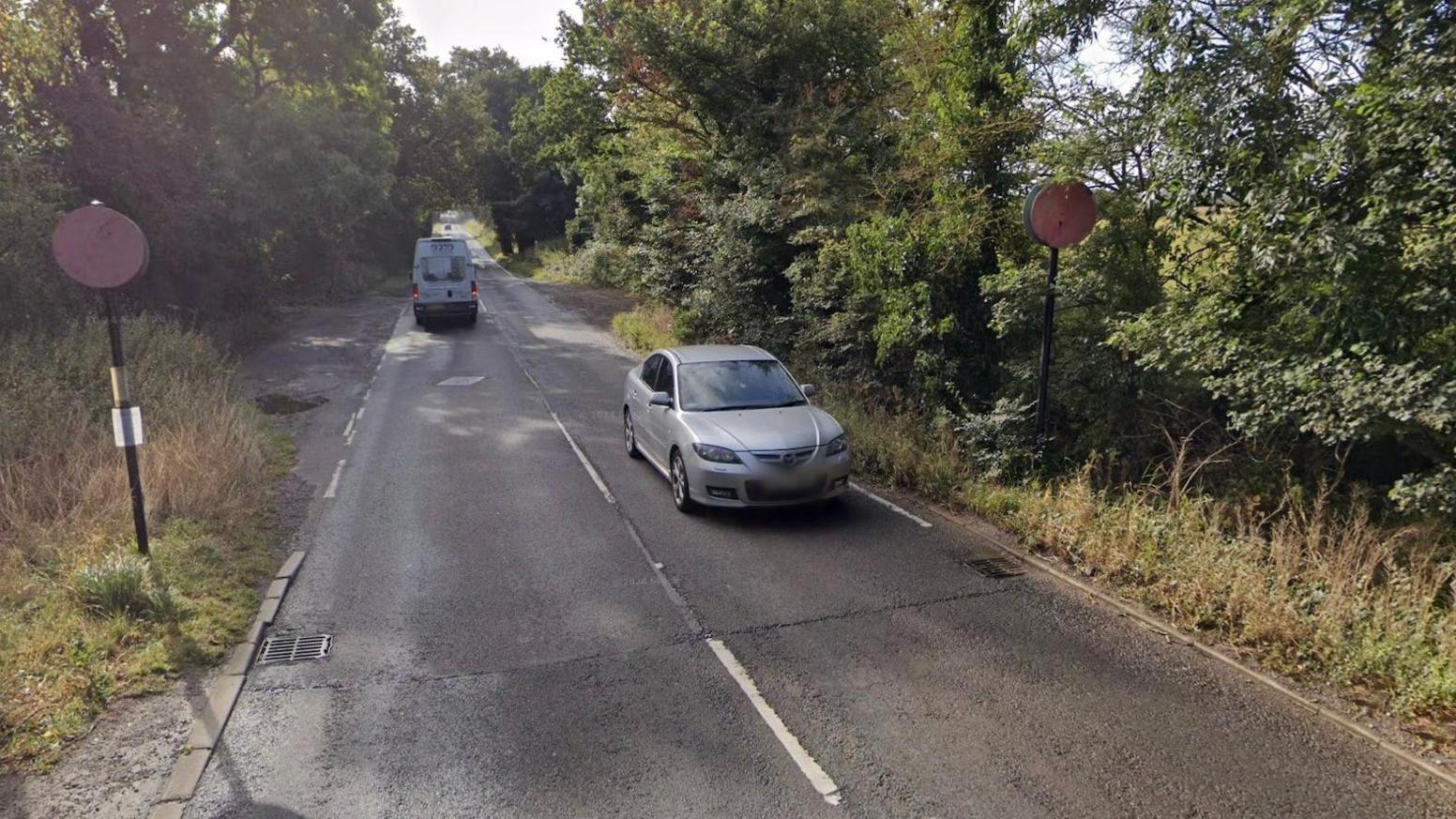 A Google street view image of a road with no pavement or footpath, showing cars driving along it in both lanes