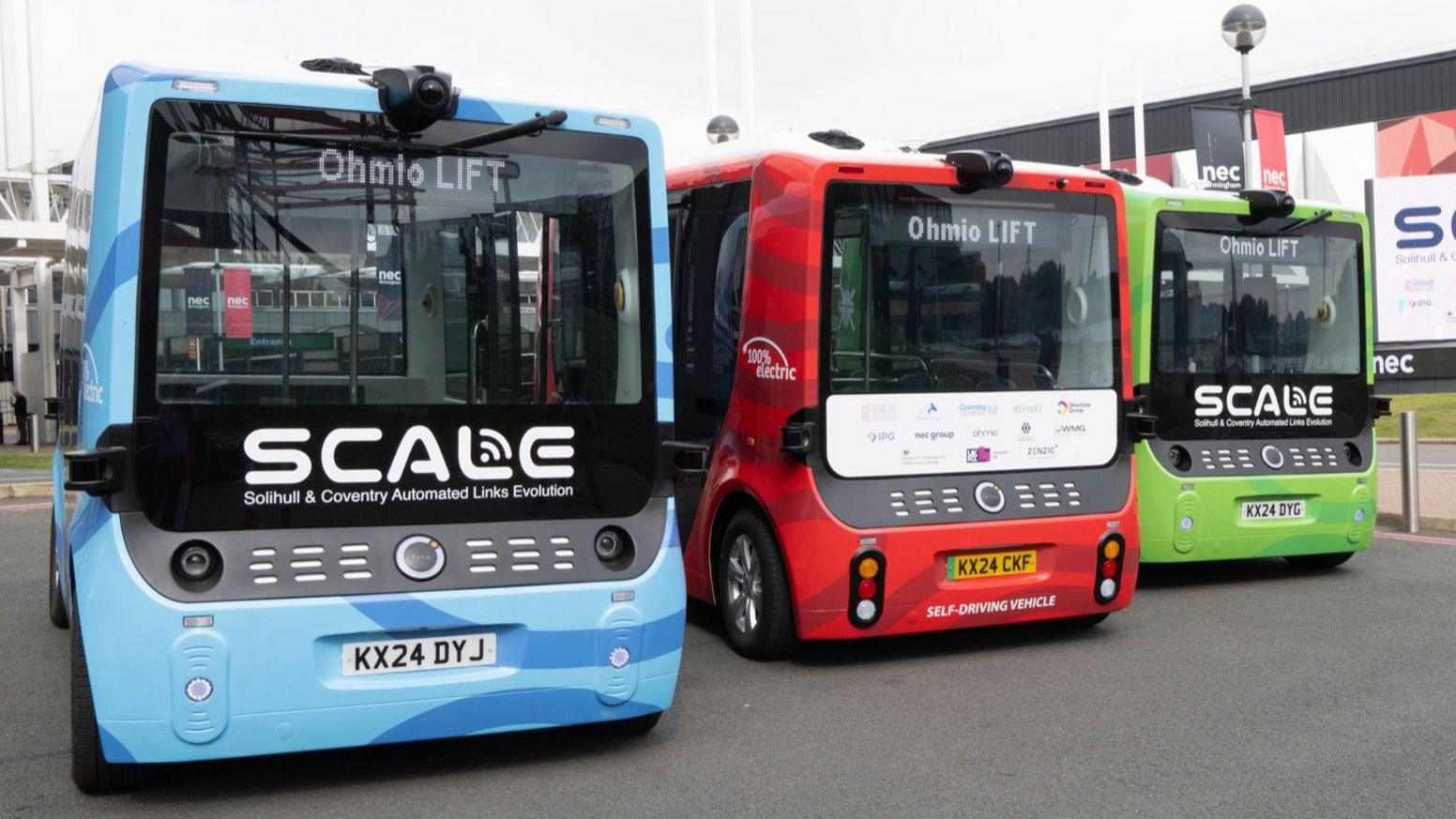 Three short, self-driving shuttle buses side by side. One is blue, one is red, and one is green.