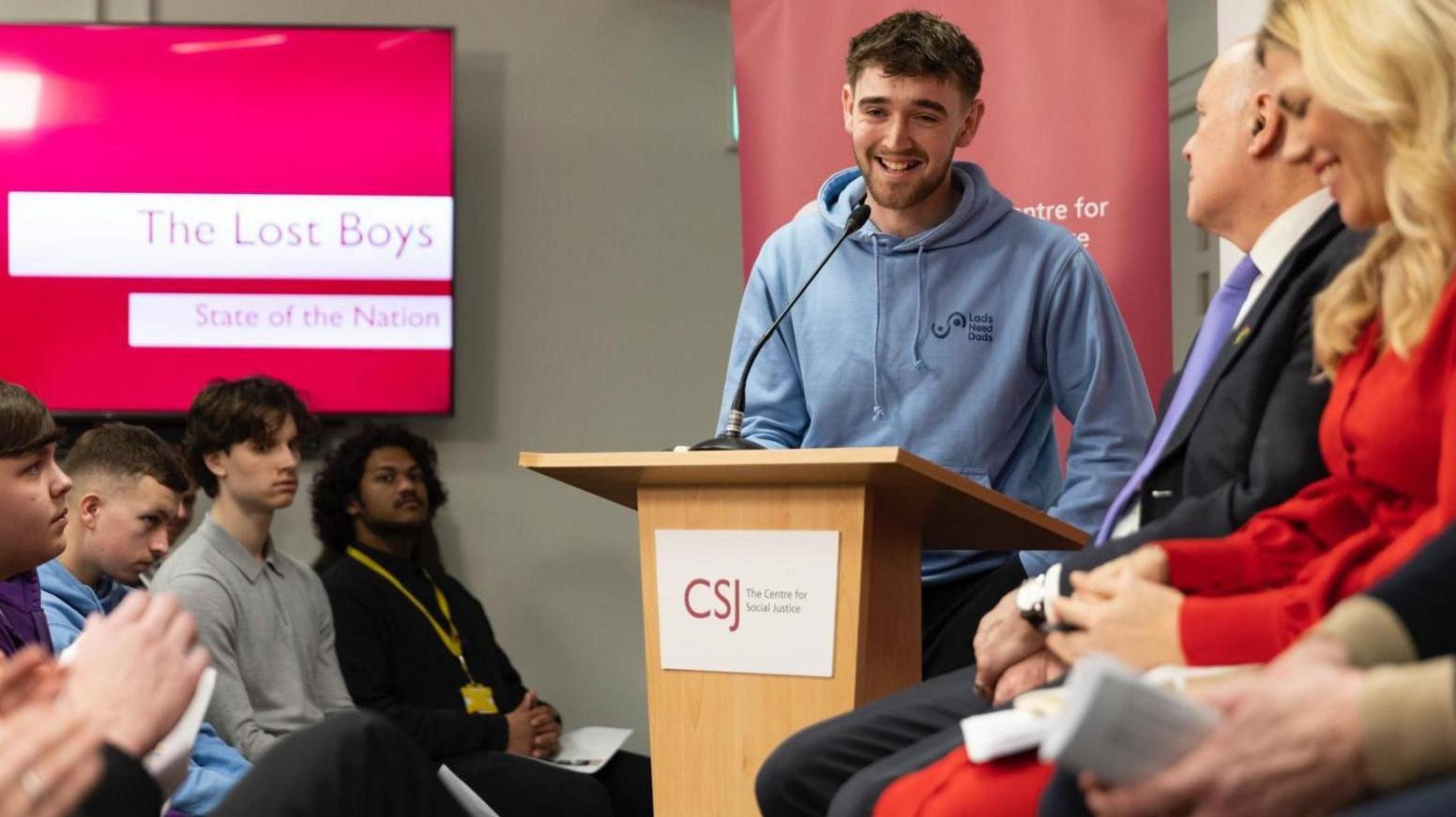 Charlie at a lecturn smiling, with people listening to him