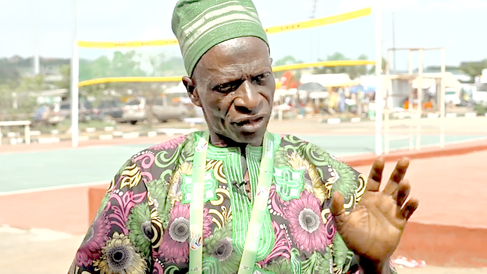 A picture of a man in his late-seventies, wearing a largely pink and green floral African fabric and a green hat raises his left hand slightly to display his palm to the camera during an interview. An Abula court can be seen in the background, with a net with yellow trim top and bottom