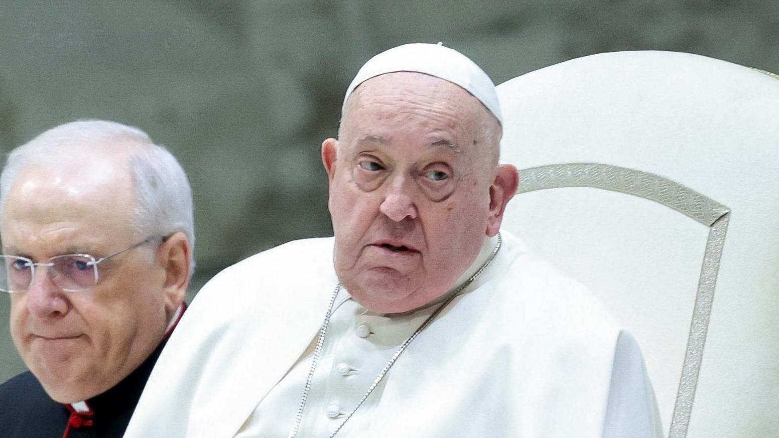 Pope Francis, wearing white robes and hat, sits on a white chair with another man, wearing black robes, next to him