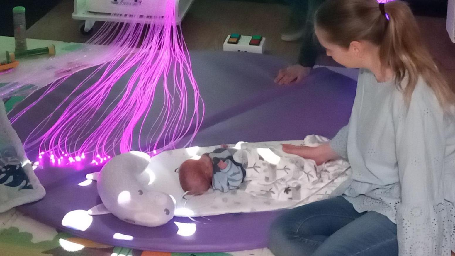 A  woman patting a new-born baby lying on an animal character mat on a colourful floor with lines of purple light nearby