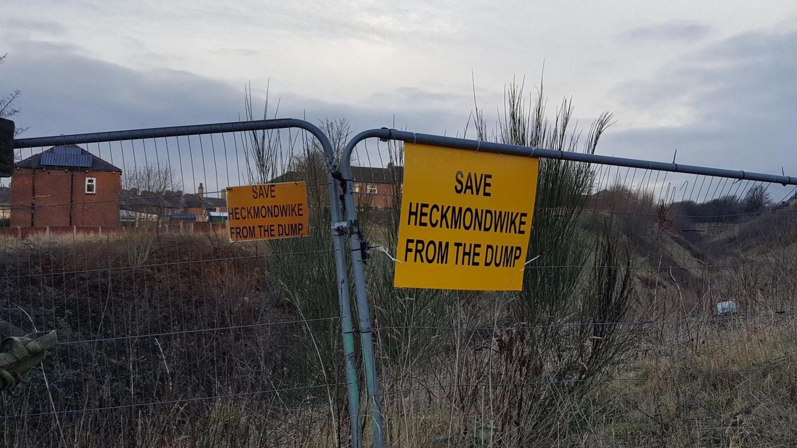 Metal fences around a disused area of grass. On them yellow signs read 'Save Heckmondwike from the dump'