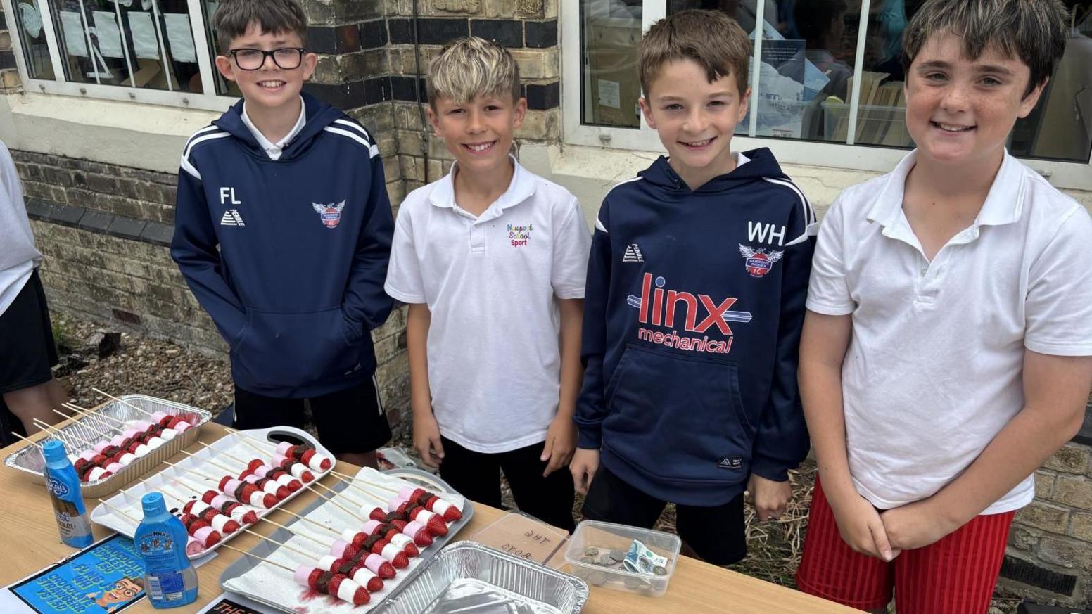 Four schoolchildren standing behind a table, two wearing blue tracksuit tops and two wearing white polo shirts. There are a number of trays on the table containing what appears to be marshmallow kebabs.