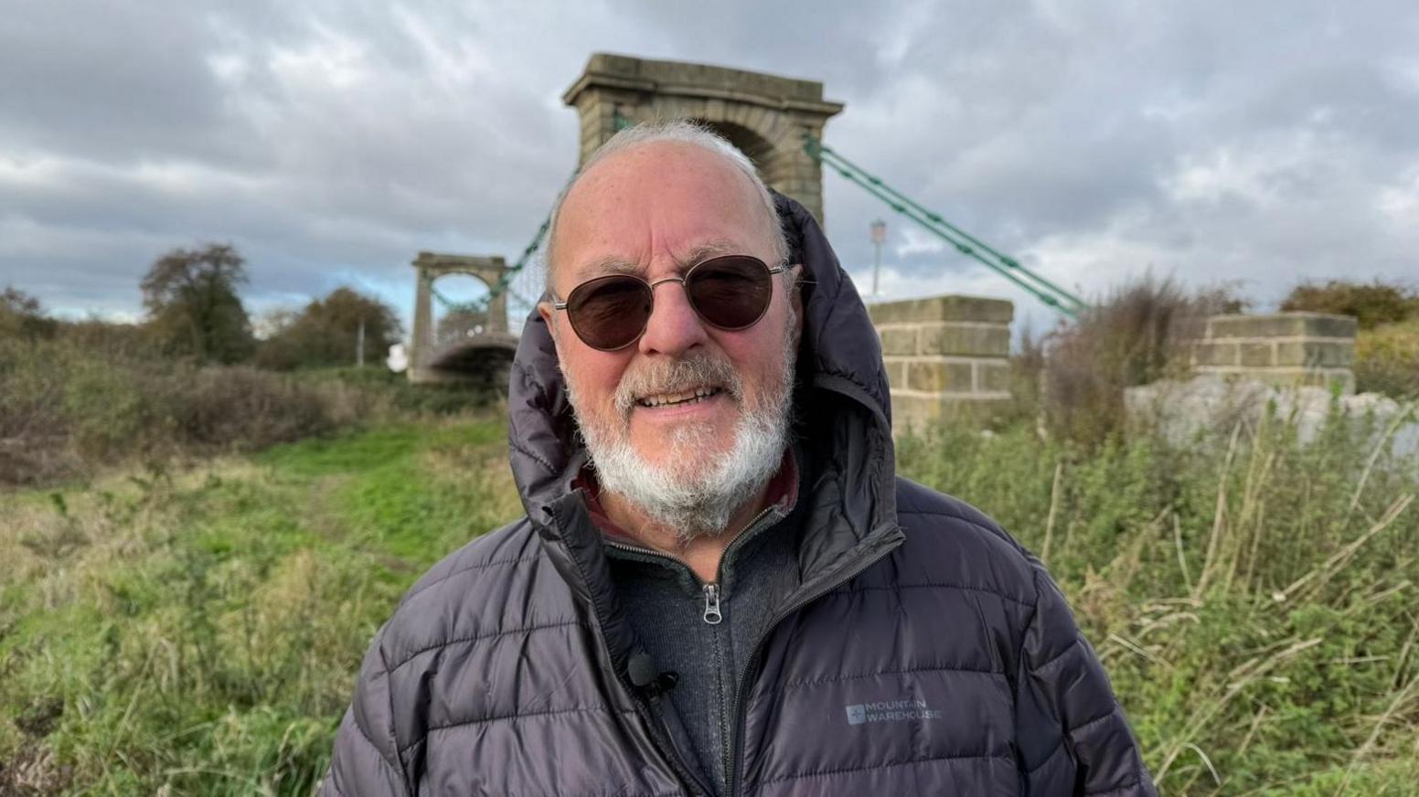 The photo shows a man looking at the camera. He's wearing a black coat with a hood up. He's also wearing sunglasses. He's standing in front of a suspension bridge, and lots of greenery.