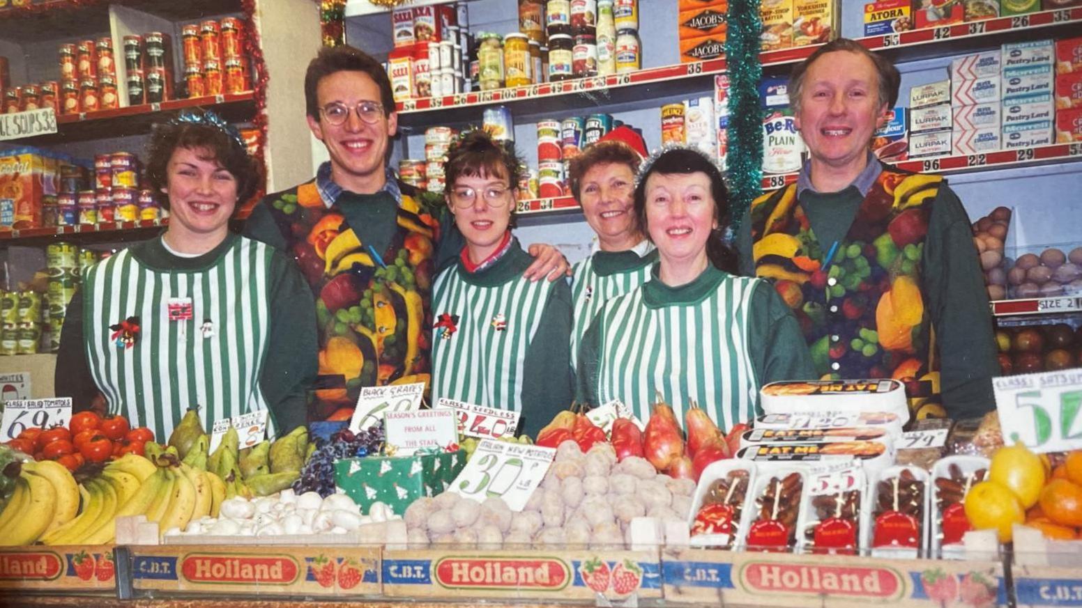 A group of staff lined up in matching uniform behind boxes of fruit and veg. Female workers are wearing outfits with vertical green and white stripes. The two men in the shot are wearing waistcoats covered in various fruits. 