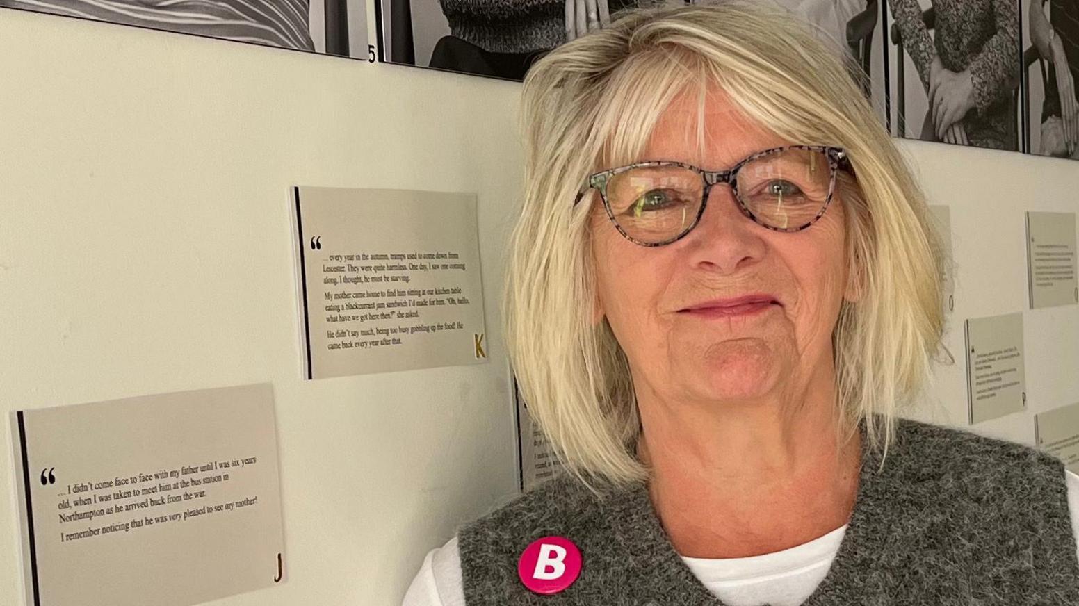 Sarah Reed stands in front of a white wall full of beautiful black and white portraits of some of the members. Sarah has white bobbed hair, black glasses a white top and grey gilet. 