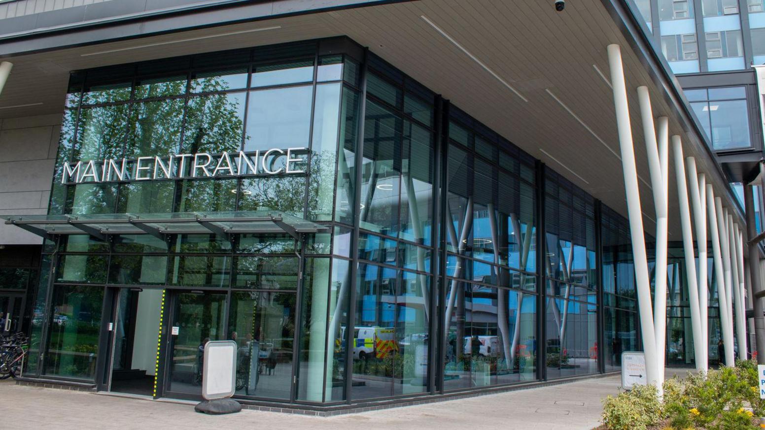 Large glass-fronted entrance to a modern building. There are white support beams and a sign which reads Main Entrance.