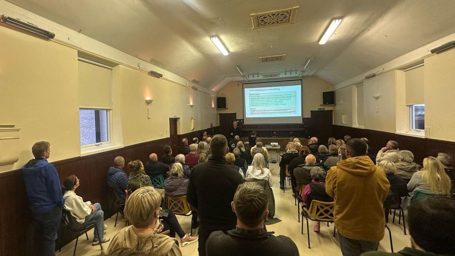 Dozens of people gathered in a community hall for a meeting. Some people are stood at the back, while those at the front are in seats. There's a large projector with text on it.