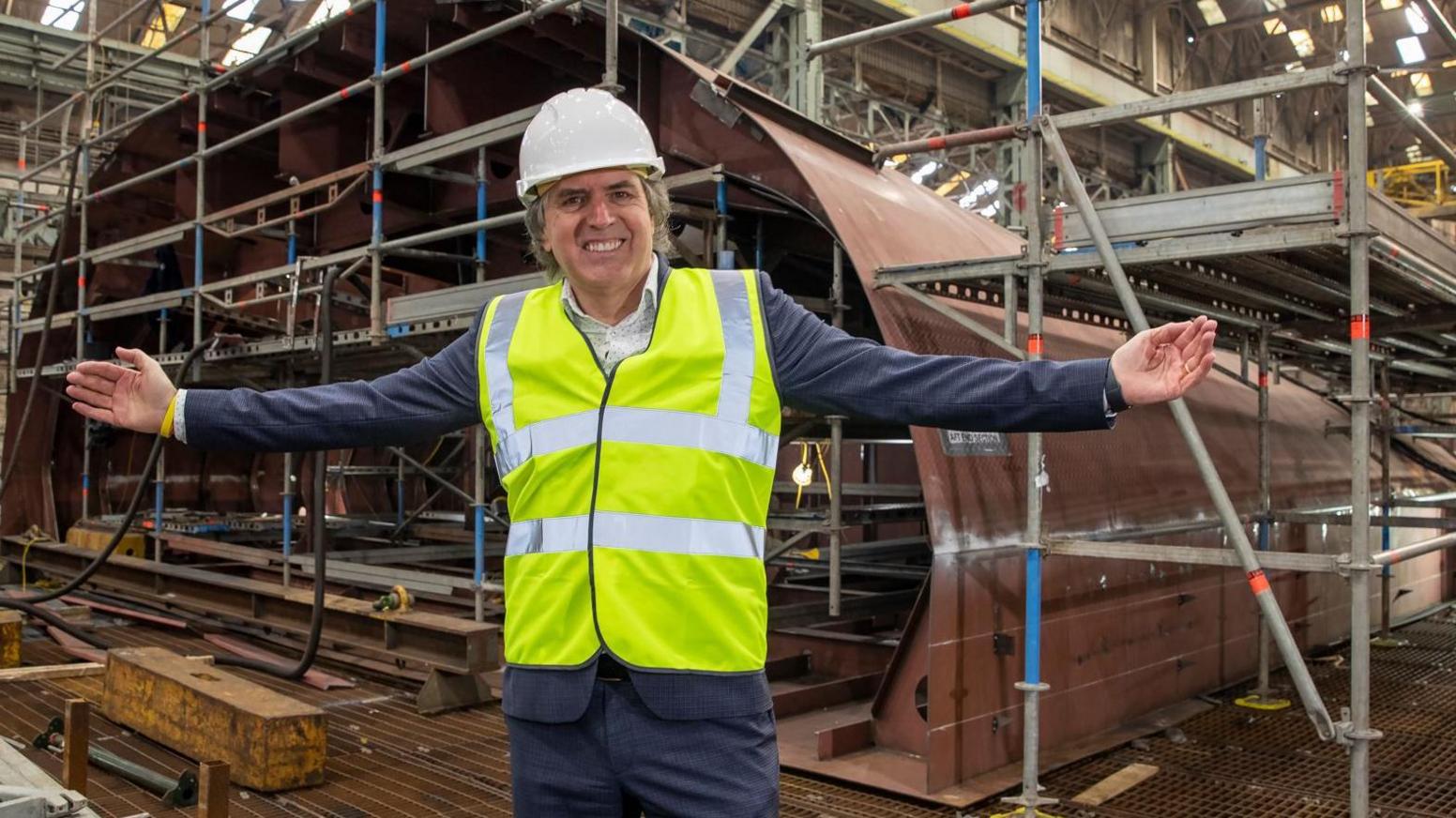 Steve Rotheram is stood next to construction of the Royal Daffodil mersey ferry. He is wearing a white hard hat and yellow high visibility vest. He is standing with his arms reaching out to the side and smiling at the camera.