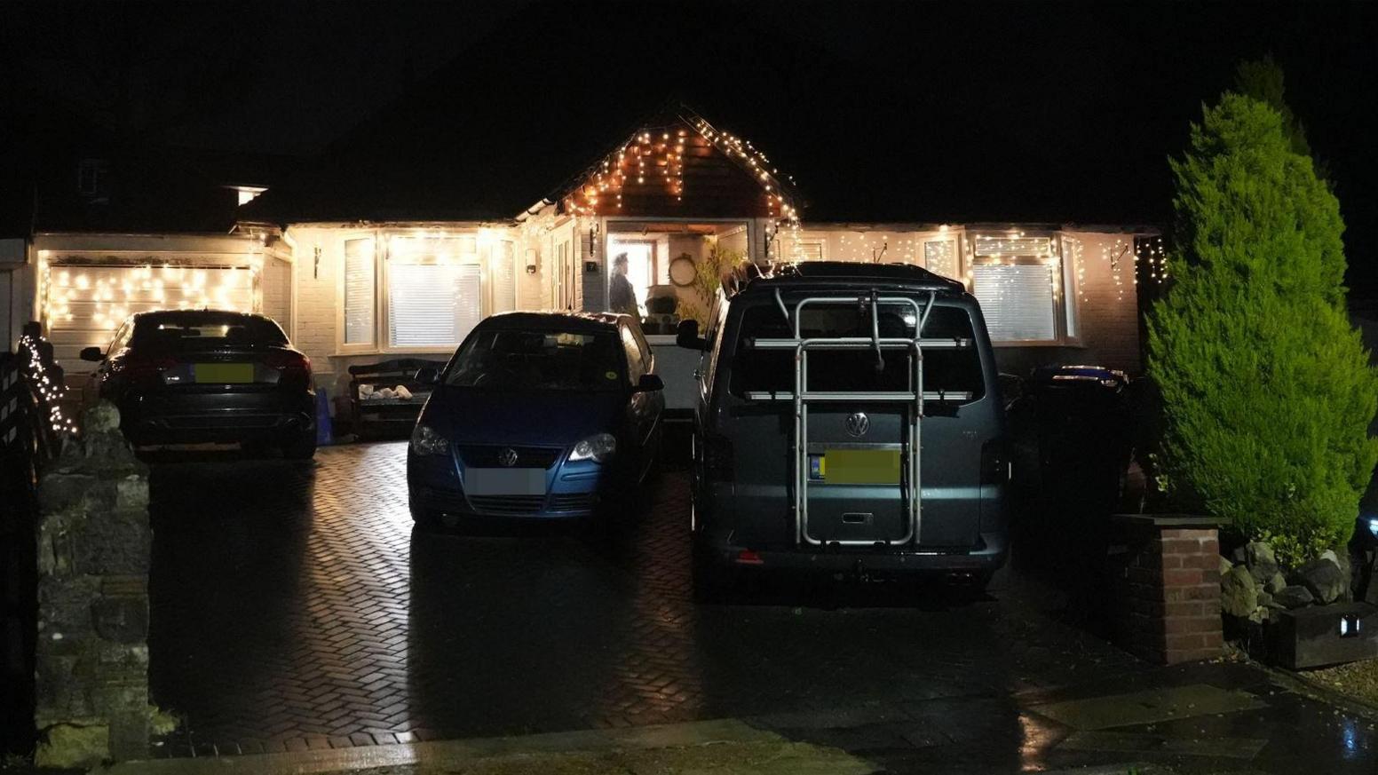 A night-time external shot of a house in East Grinstead festooned in Christmas lights with three cars outside which is the scene of an alleged murder