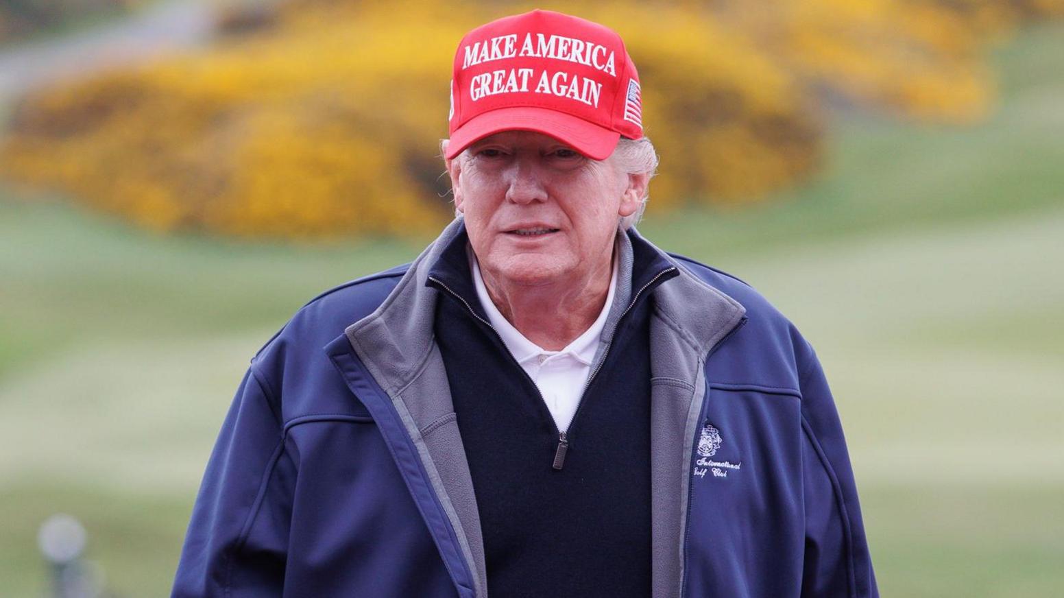 Donald Trump in red Make America Great Again golf baseball hat, dark blue Trump Turnberry branded fleece jacket, white polo shirt and blue jumper, is standing in front of a tee at Turnberry. The course can be seen behind him.