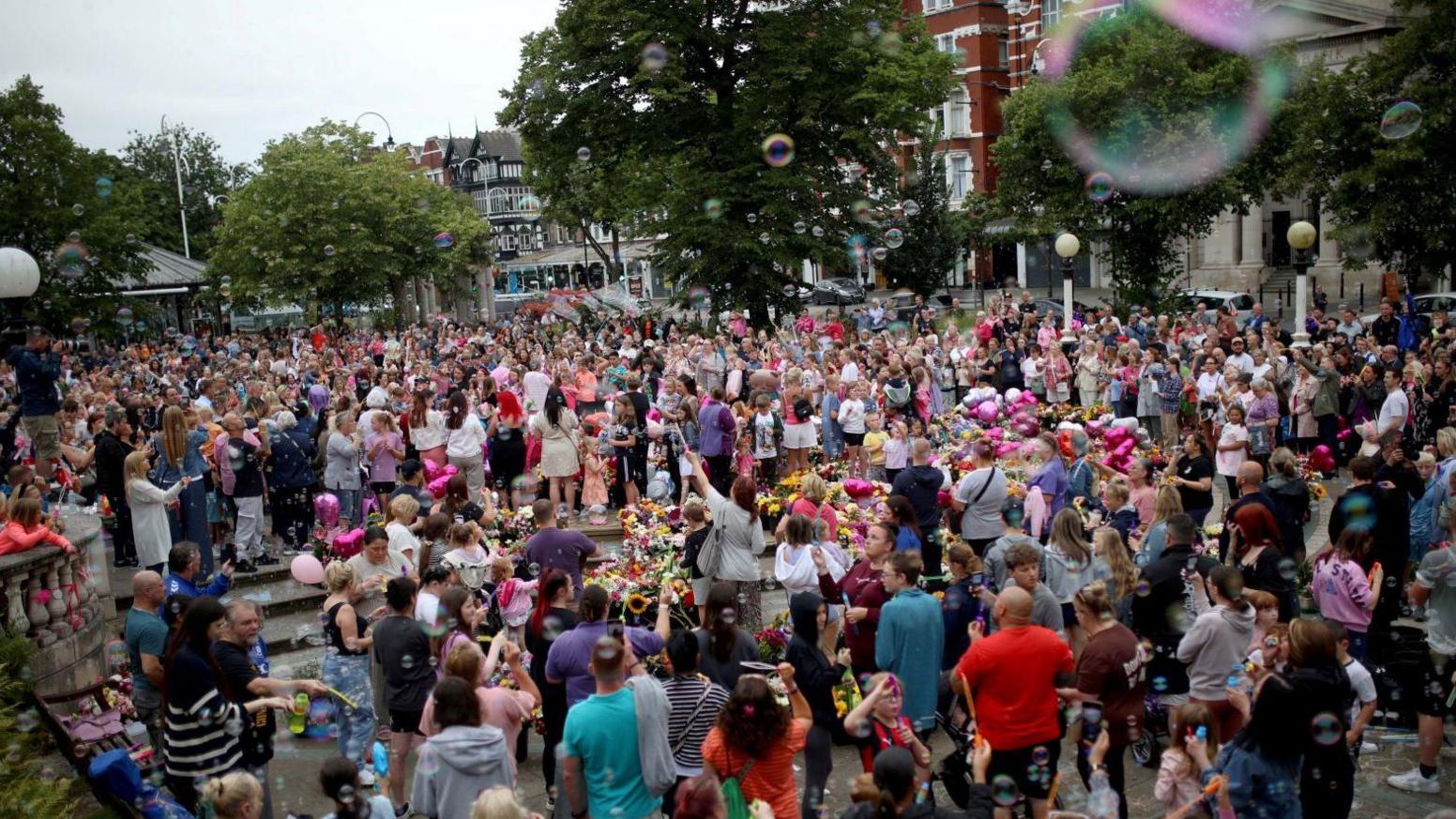 Crowds gather in Southport town centre. There are floral tributes and pink balloons and many people are blowing bubbles up towards the sky, which is relatively cloudy and grey.