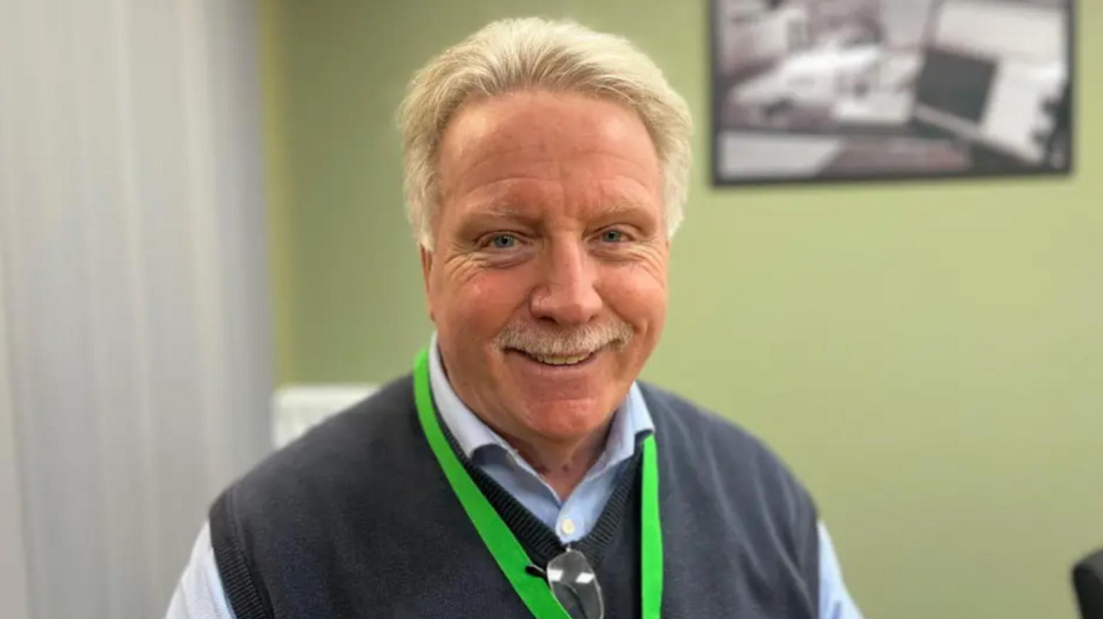 A man wearing a light blue shirt with a dark blue knitted vest and a green lanyard stands, smiling, in front of a light green wall.
