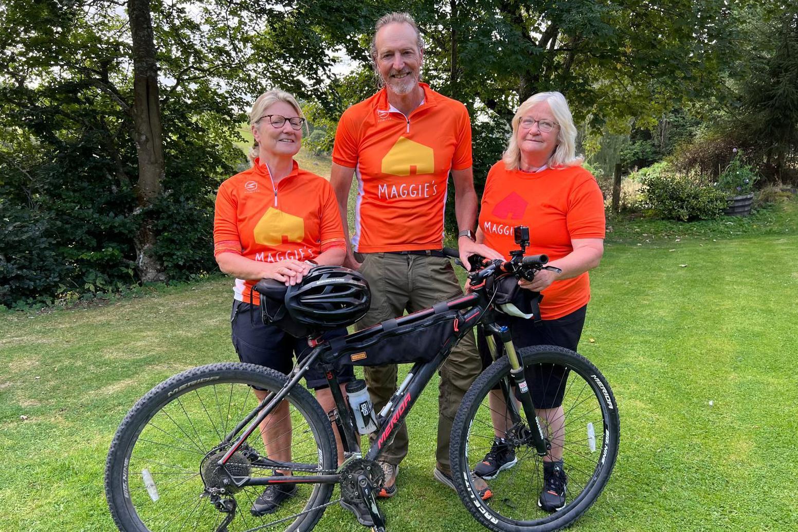 Rona, Craig and Sophie as they are today. They are standing in a garde with a black mountain bike. They are wearing orange cycling tops with the word "Maggie's" in white lettering.