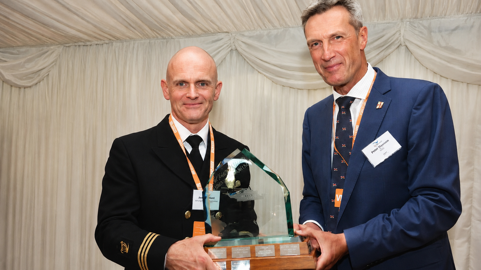 Two men are looking at the camera smiling while holding a glass award. The man on the left is bald and dressed in a uniform, the man on the right has grey hair and is dressed in a dark blue suit. 