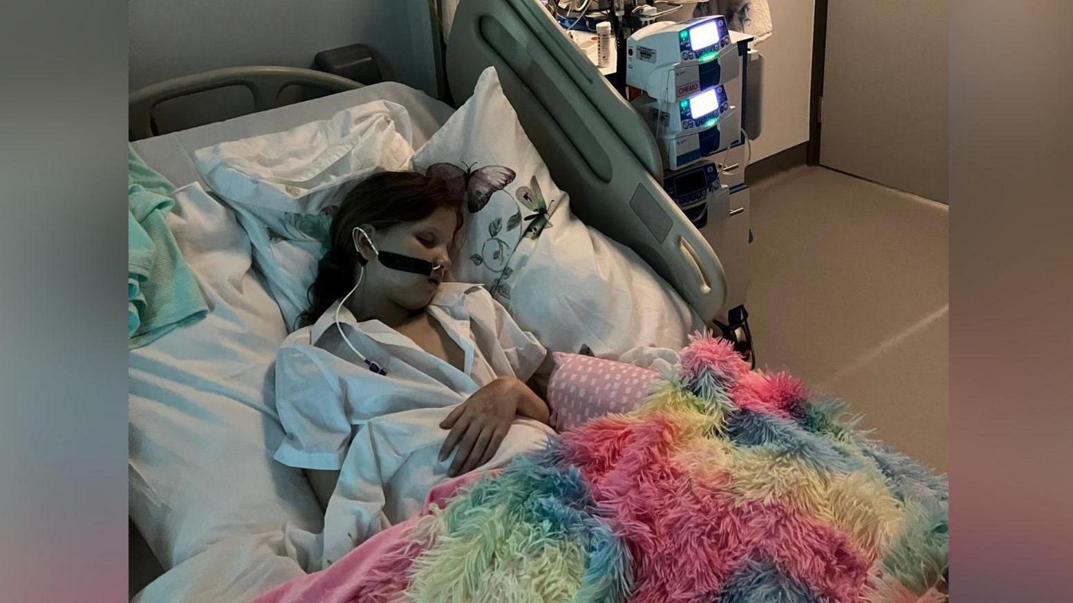 Llun o Kathryn yn cysgu yn ei gwely yn yr ysbyty. Mae'n gwisgo pyjamas gwyn ac mae ganddi flanced liwgar.