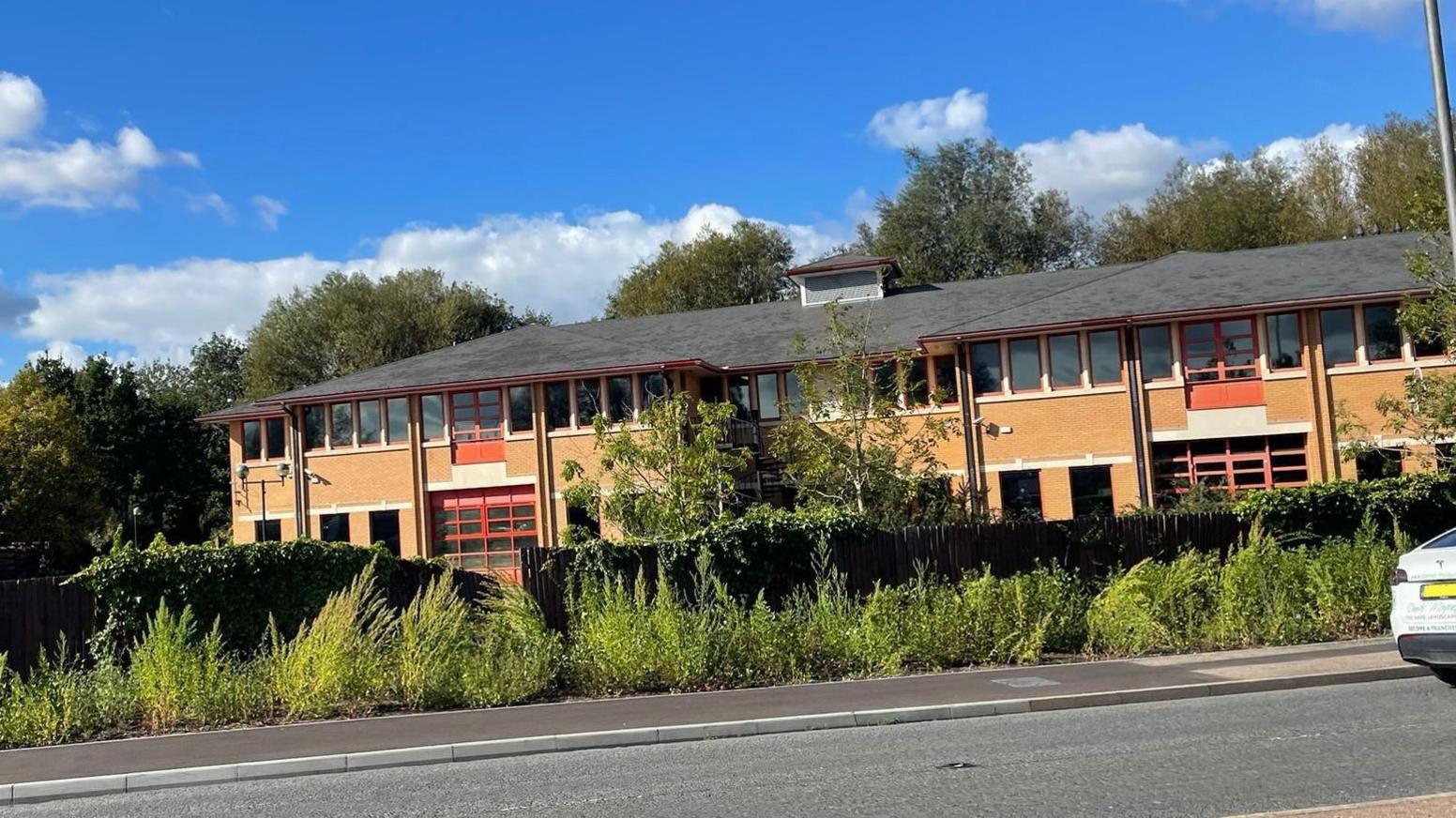 A two-storey brick building with red window frames surrounded by vegetation and next to a main road.