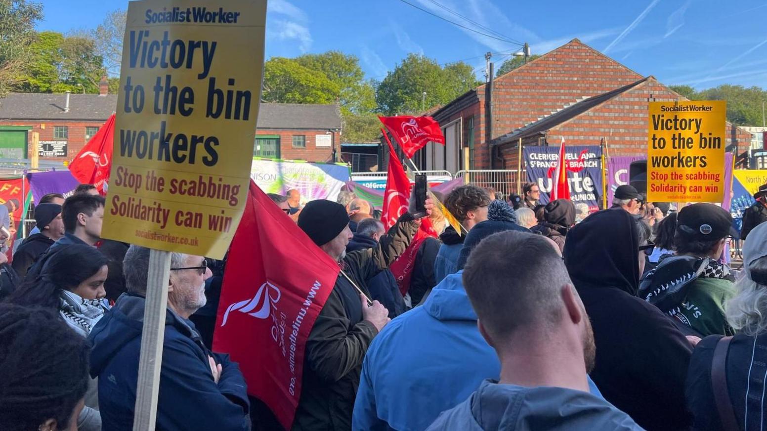 The backs of demonstrators who are holding up several signs including a yellow one from the Socialist Worker which says "Victory to the bin workers". There are also several red signs. Some of the protesters are wearing caps or beanie hats. Many are also wearing fleece jackets or coats.