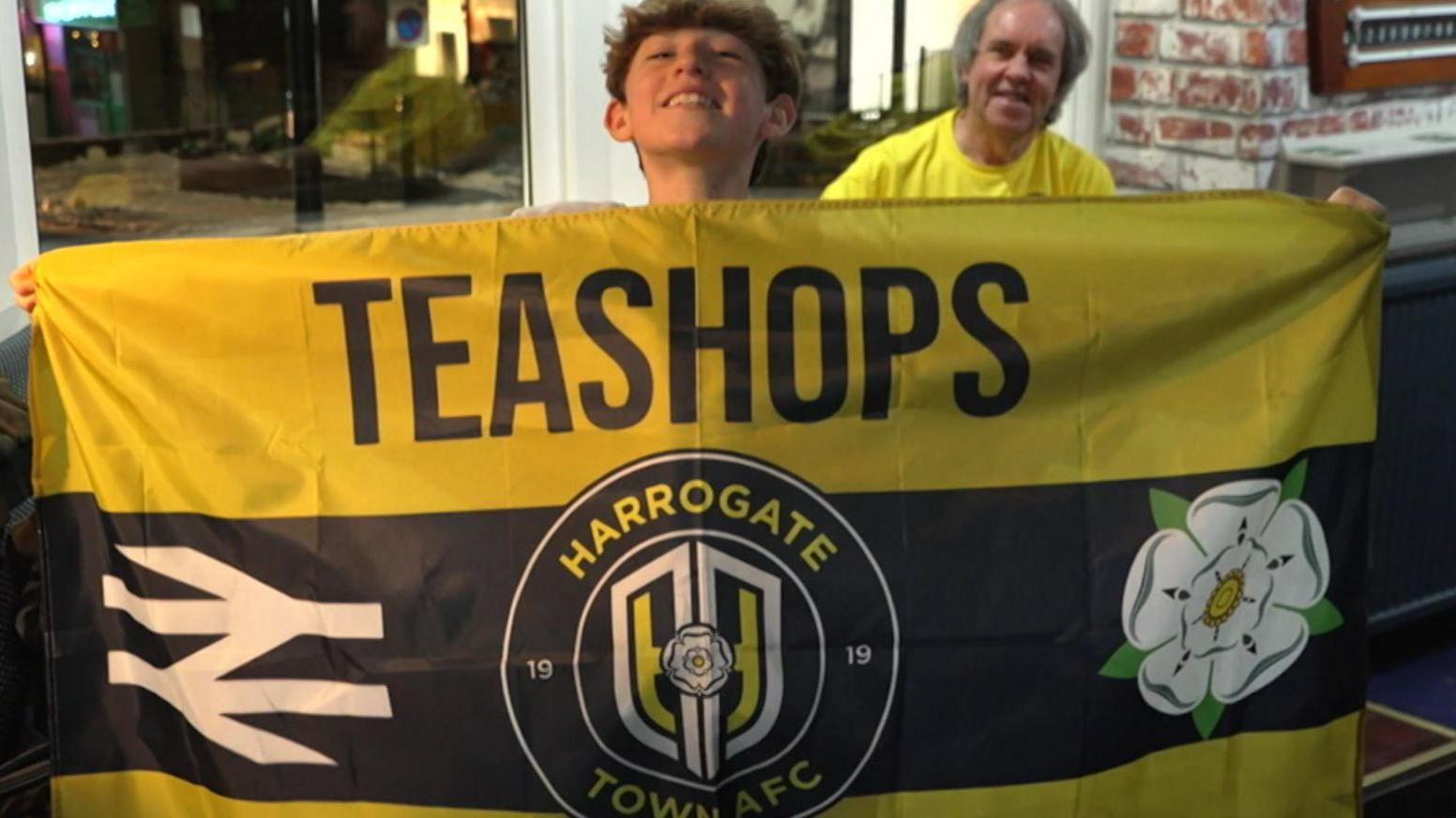 A young Harrogate Town AFC supporter holds up a yellow flag with "teashops" written on it in bold black letters.