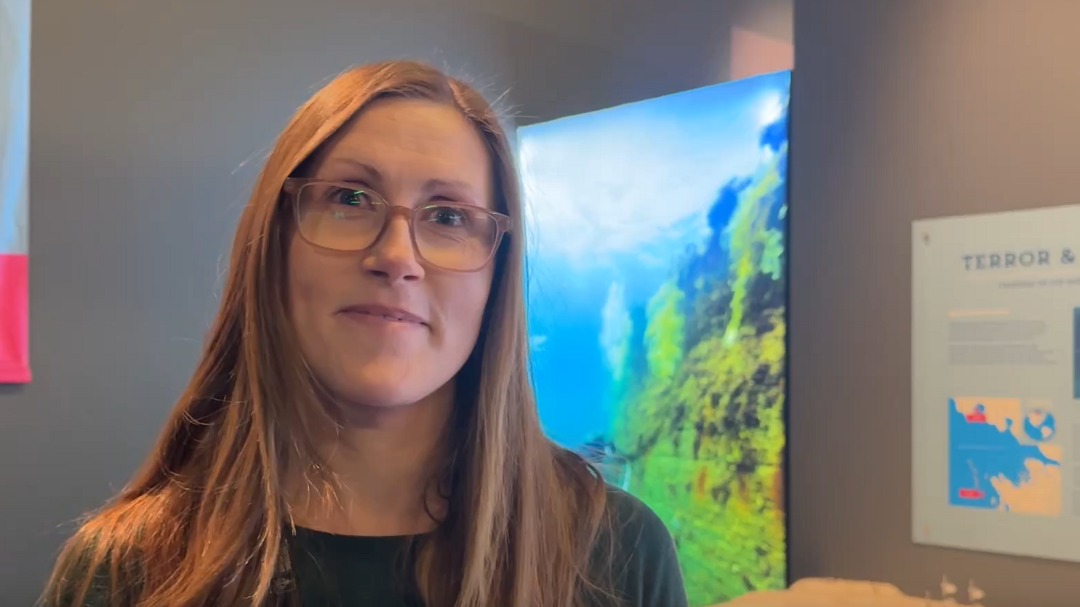 A woman with brown hair and glasses stood in a room with a sea bed image behind her