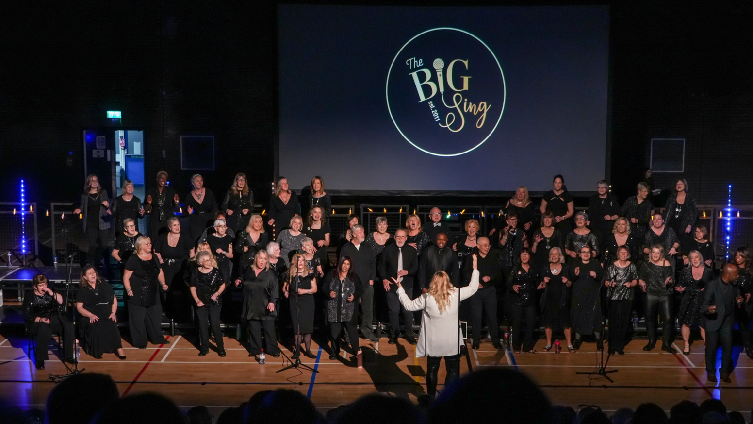 A large choir dressed in black clothing performing for an audience in front of the BIG Sing logo. A woman in white is conducting the choir.