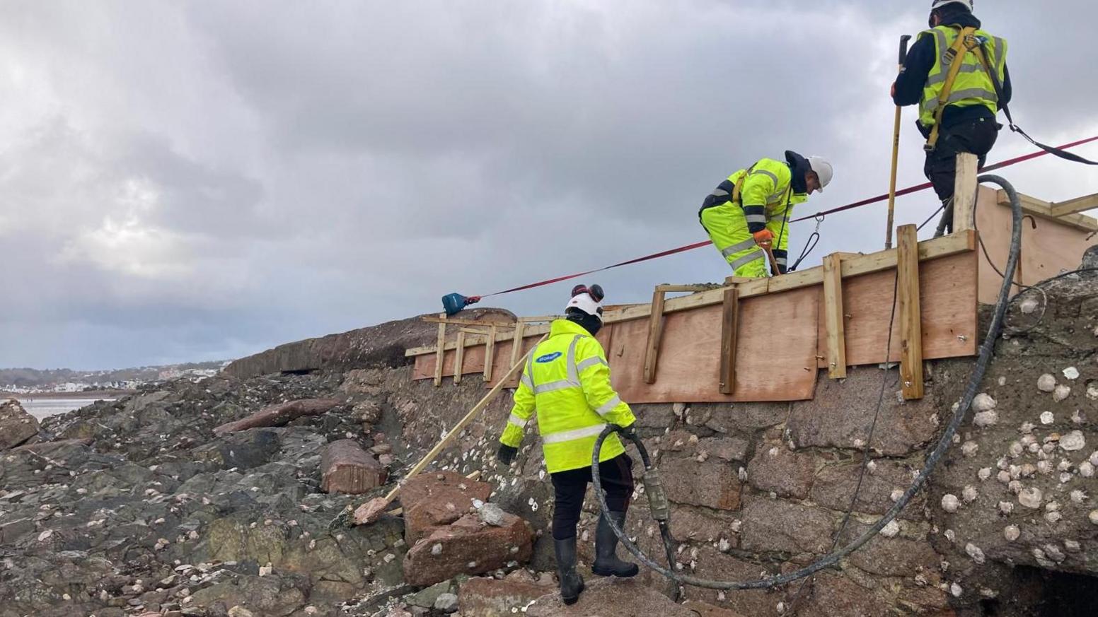 A group of workers working on a damaged seawall. There is a wooden structure around the wall with a two men working on it. In front of the wall is rocks covered in seaweed and shells.