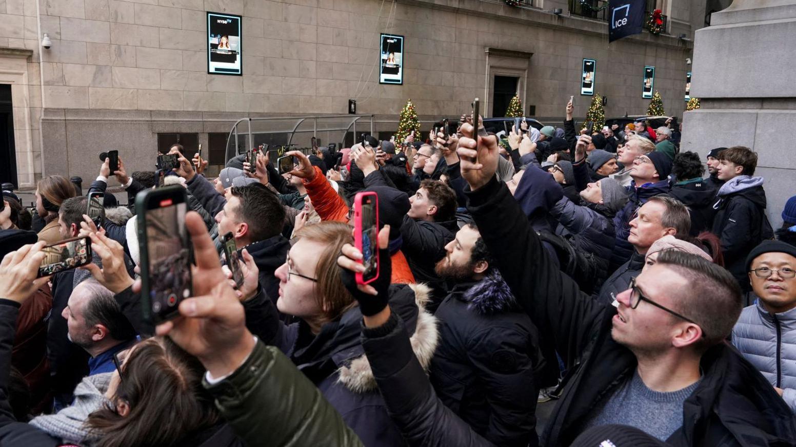 People crowded and in coats gather outside the NYSE with their phones in the air to watch US President-elect Donald Trump. Trump is not pictured, just a large packed crowd 