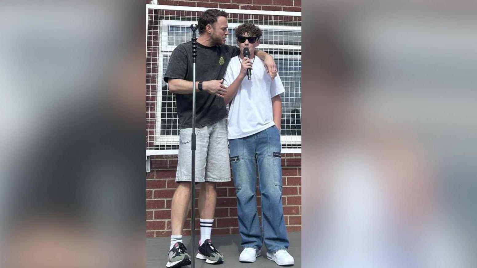 Olly Murs has a pair of grey shorts and a darker grey t-shirt on with his arm around Charlie who is singing into a mic on stage.